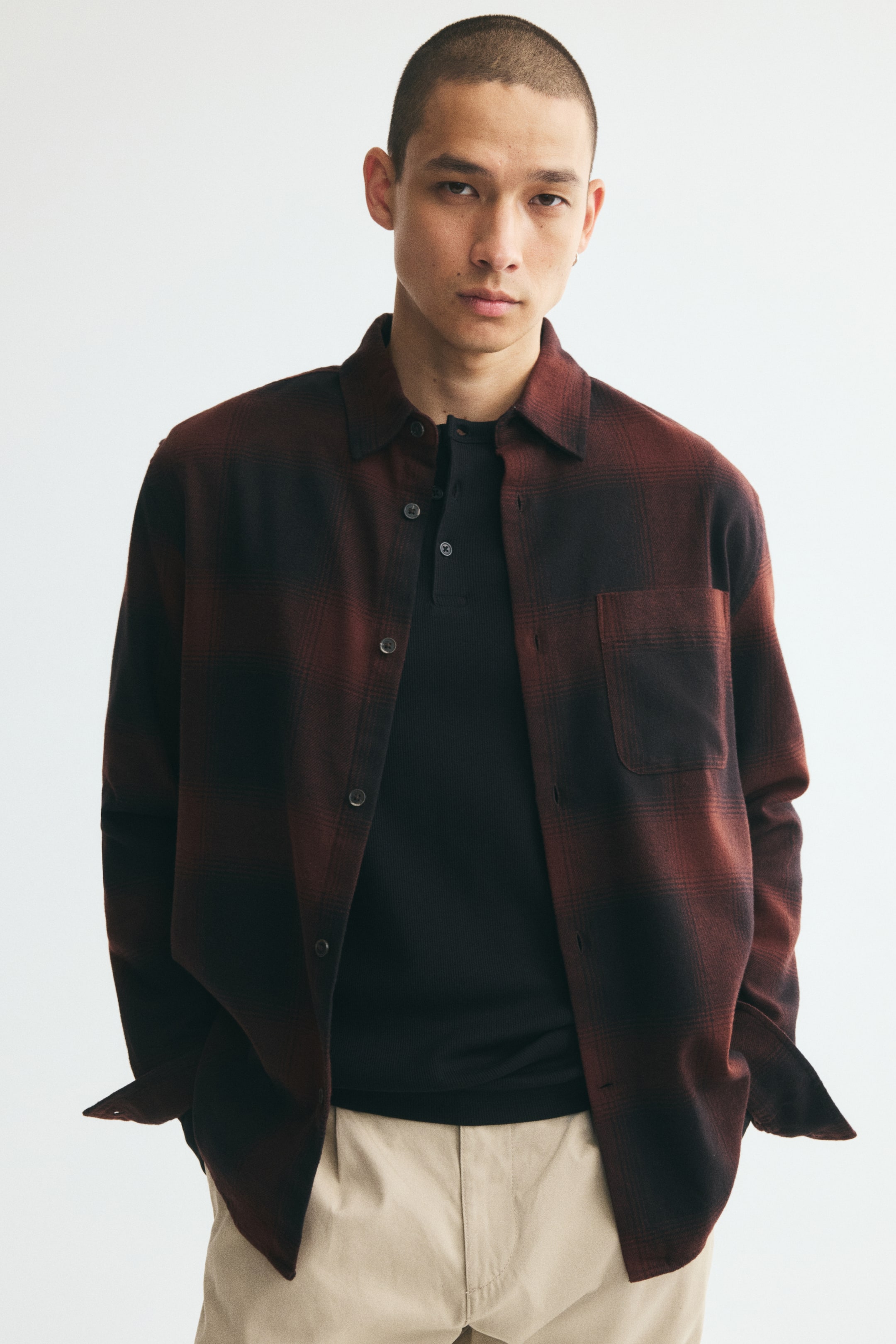 Agrandir l'image: Une chemise en flanelle à coupe ample, à carreaux rouge foncé et noirs, est portée ouverte par le mannequin sur un t-shirt henley noir et un pantalon beige. La chemise présente un col rabattu, une seule poche poitrine ouverte et des manches longues avec poignets non boutonnés.