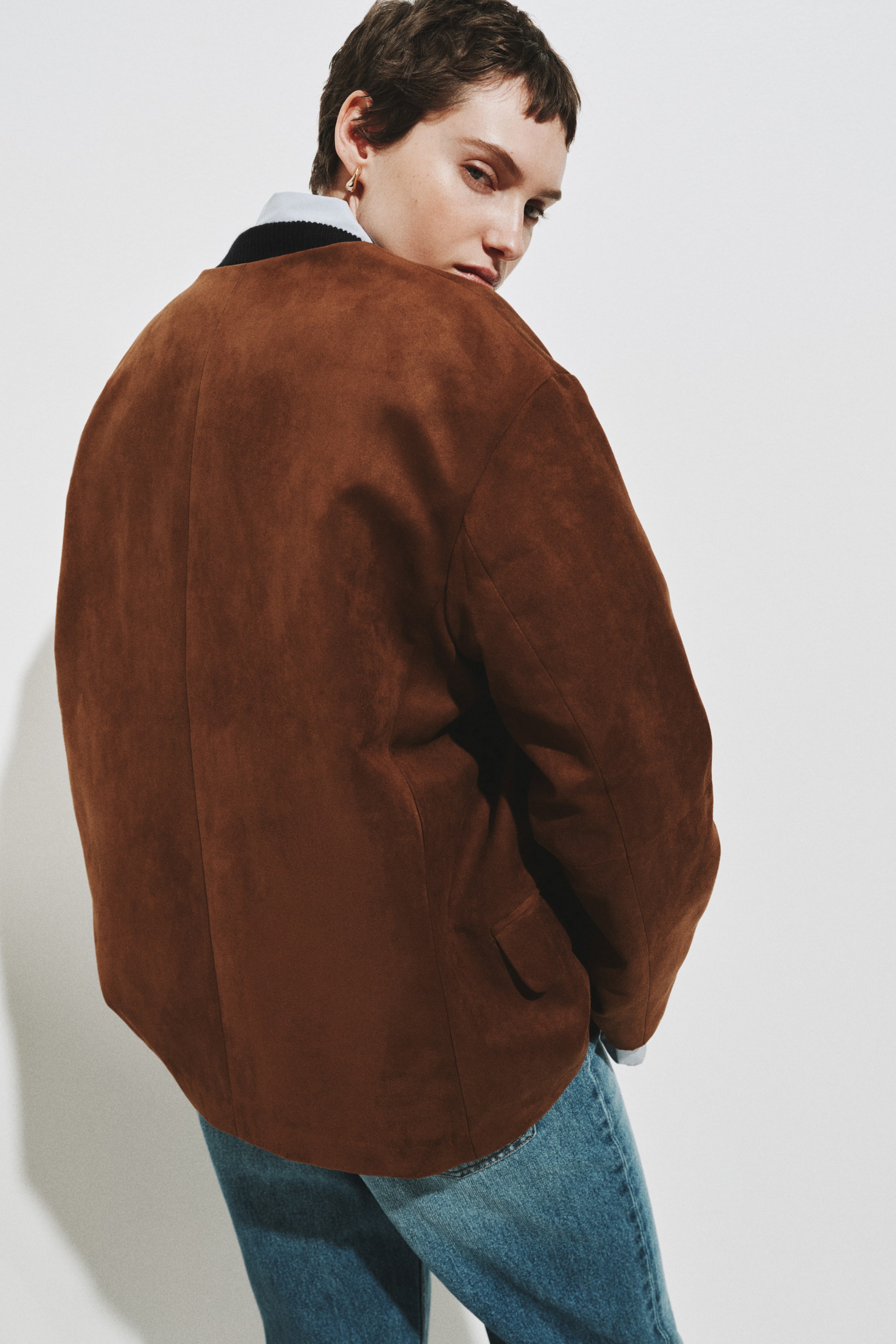 Visualizza immagine più grande: Una giacca oversize marrone mostra la parte posteriore, con una texture morbida dall'effetto scamosciato e un orlo arrotondato. Una tasca a patta è visibile sul lato destro e un colletto di camicia azzurro chiaro si intravede all'altezza del collo, insieme ai jeans blu sotto.