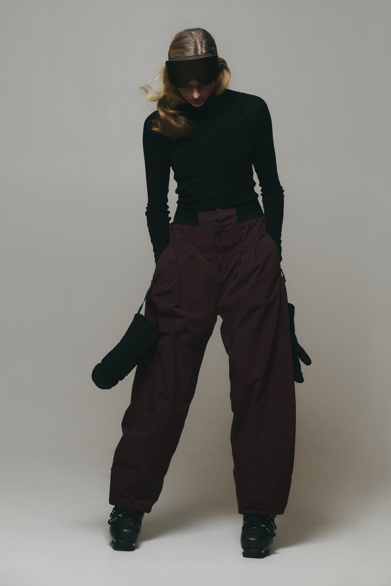 Burgundy high-waisted ski trousers with a wide-leg silhouette and front pleating are being worn by the model. The trousers are styled with a fitted black polo neck, black ski goggles, black ski boots, and black mittens hanging from the wrists. - 1