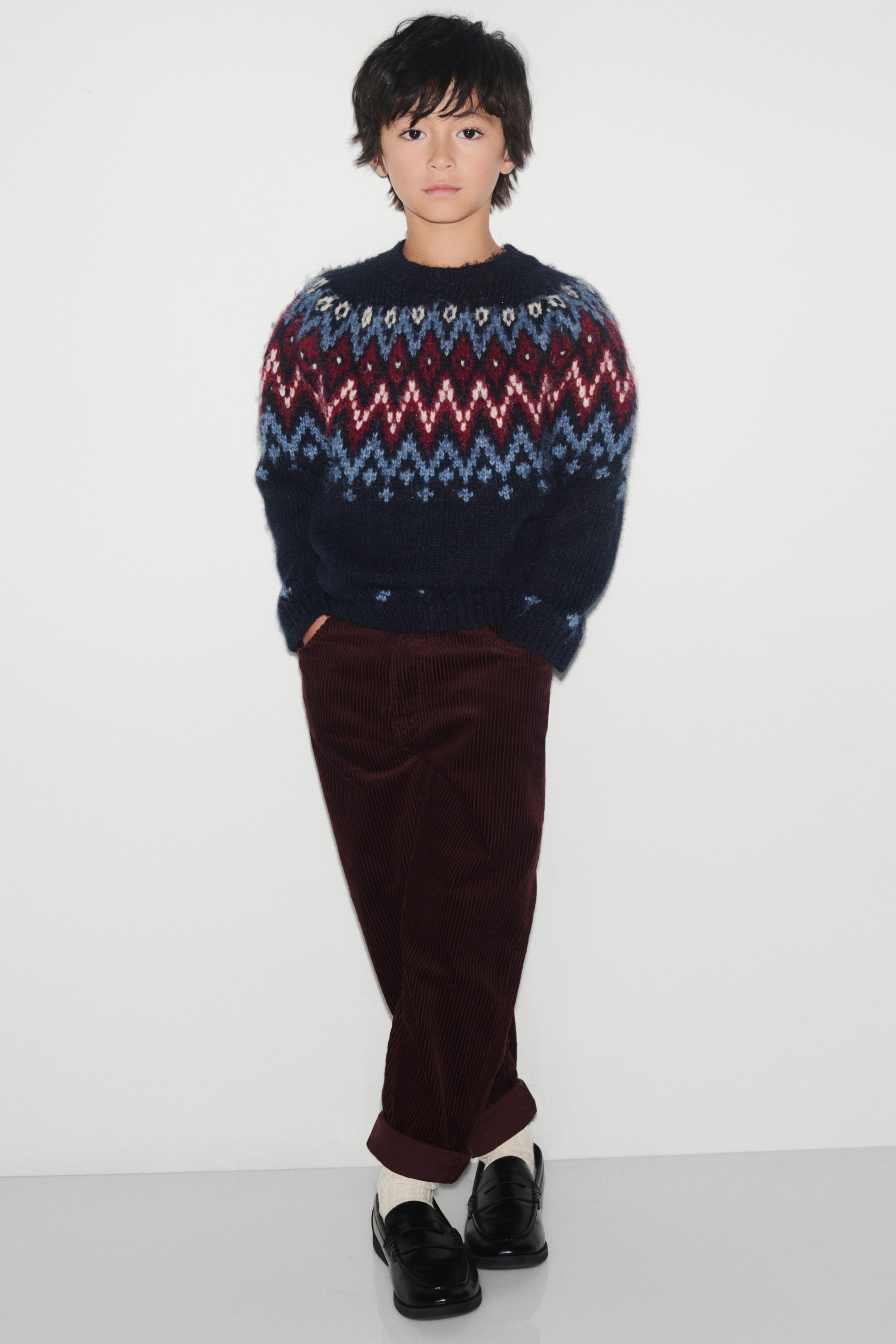 View larger image: A relaxed-fit sweater in soft navy blue jacquard knit is shown from the front, featuring a round rib-trimmed neckline and long raglan sleeves. The sweater showcases a geometric knit pattern in shades of blue, burgundy, and white across the upper chest and shoulders, and is finished with ribbed cuffs and a ribbed hem.