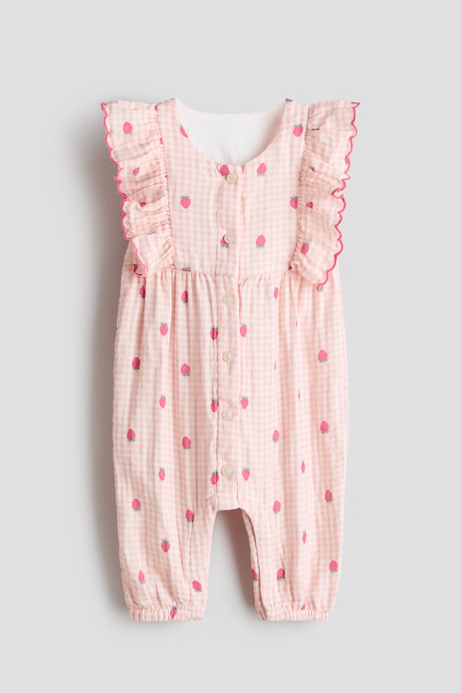 A frilled muslin romper suit is presenting its front view, and it is featuring a pink and white gingham check pattern with scattered red strawberry motifs. Frilled, sleeveless shoulders are detailing the top, and they are edged with pink scallops. A full-length front button placket is running down the center, and the suit is gathering at the waist and at the ankles of its long legs. - 1