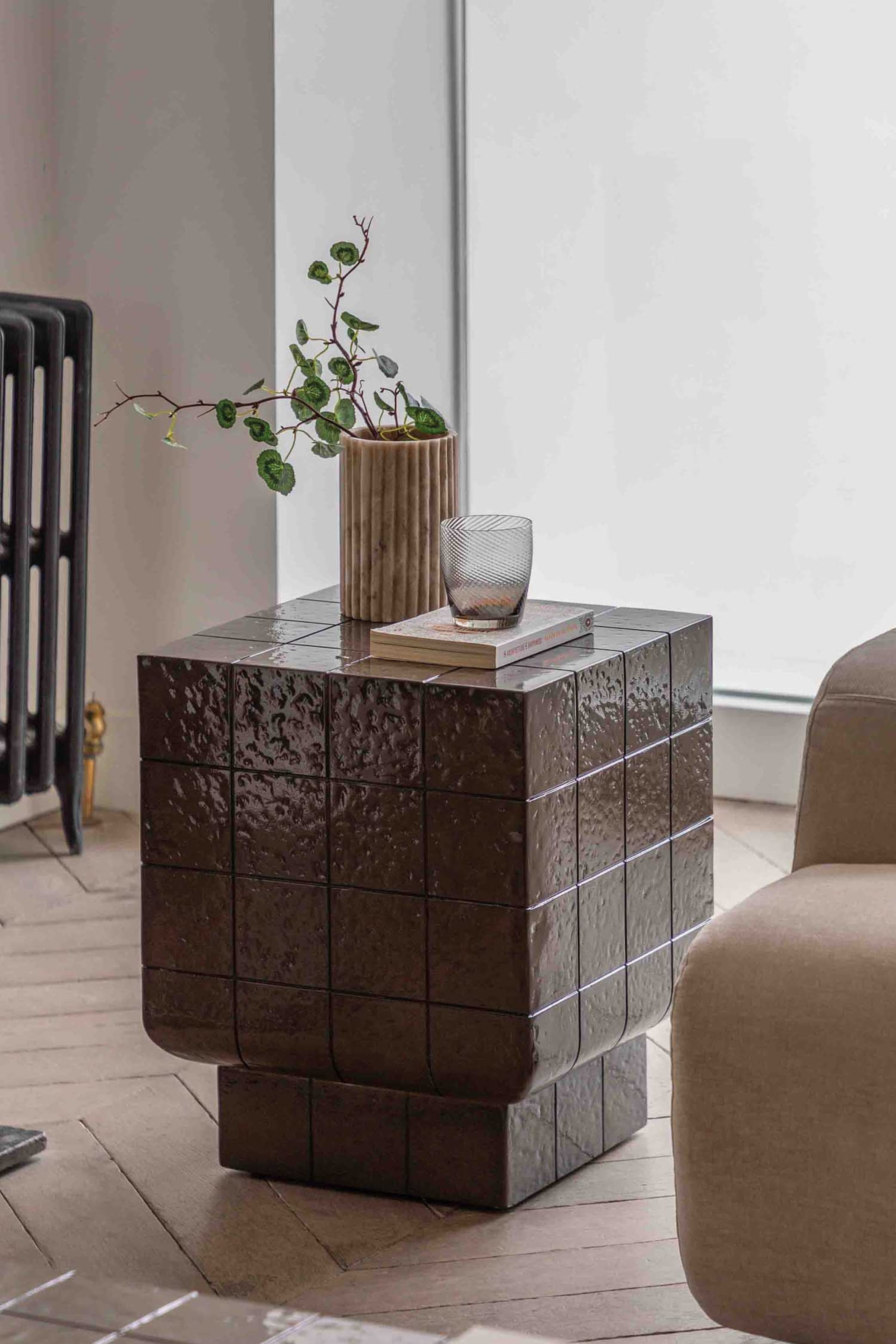 View larger image: A dark brown, cube-shaped side table featuring a glossy, textured tiled surface with visible grout lines, showcasing a smaller, recessed base and softly rounded bottom edges on its main body. A plant in a ribbed vase, a book, and a clear glass rest on its top surface.