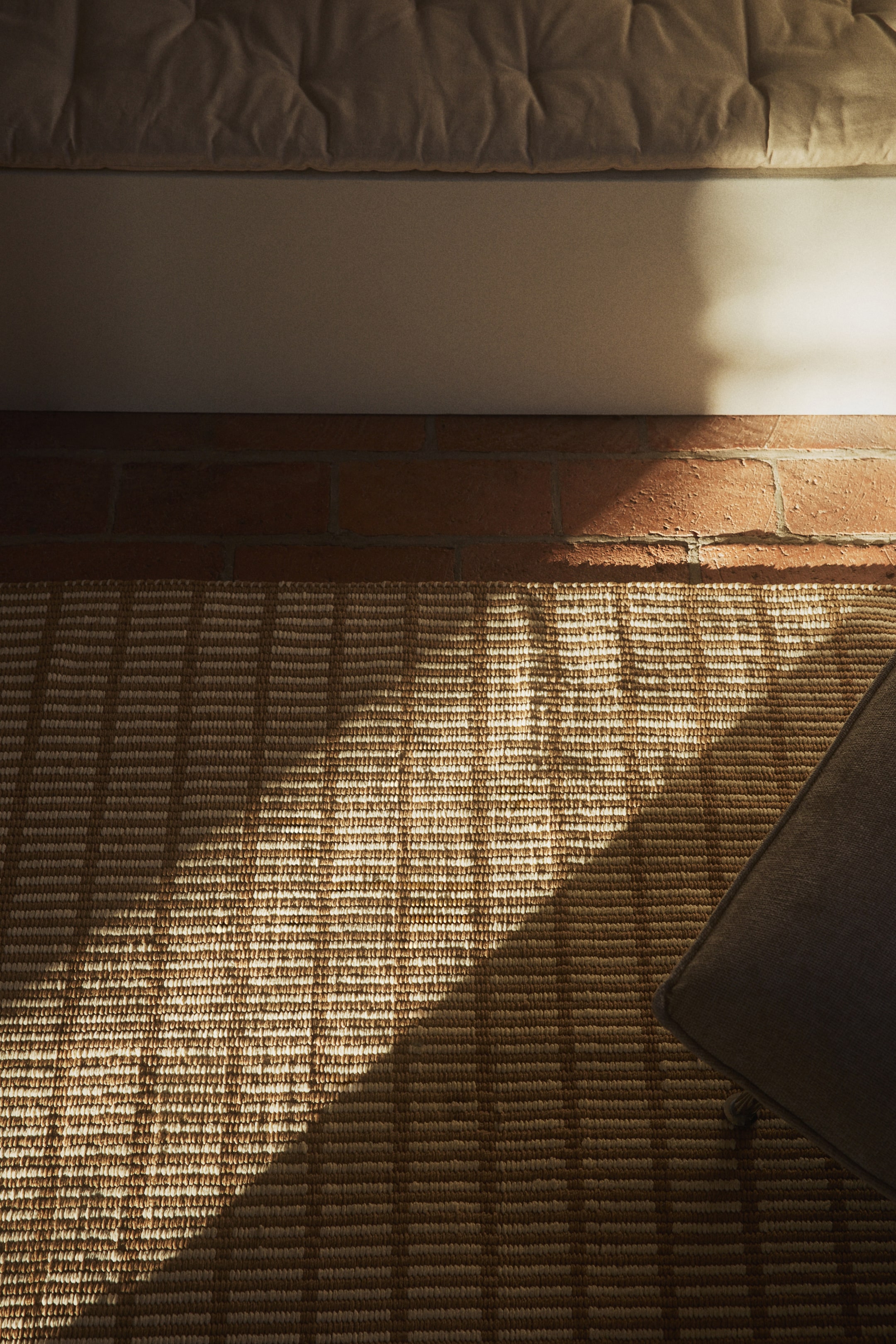 Visa större bild: En vävd matta visar ett geometriskt mönster av växlande ljusa och mörka rektangulära block arrangerade i horisontella rader och vertikala kolumner över dess yta. Den ligger platt, med en diagonal solstråle som lyser upp dess övre vänstra del, och en mörk kudde vilar i dess nedre högra hörn.