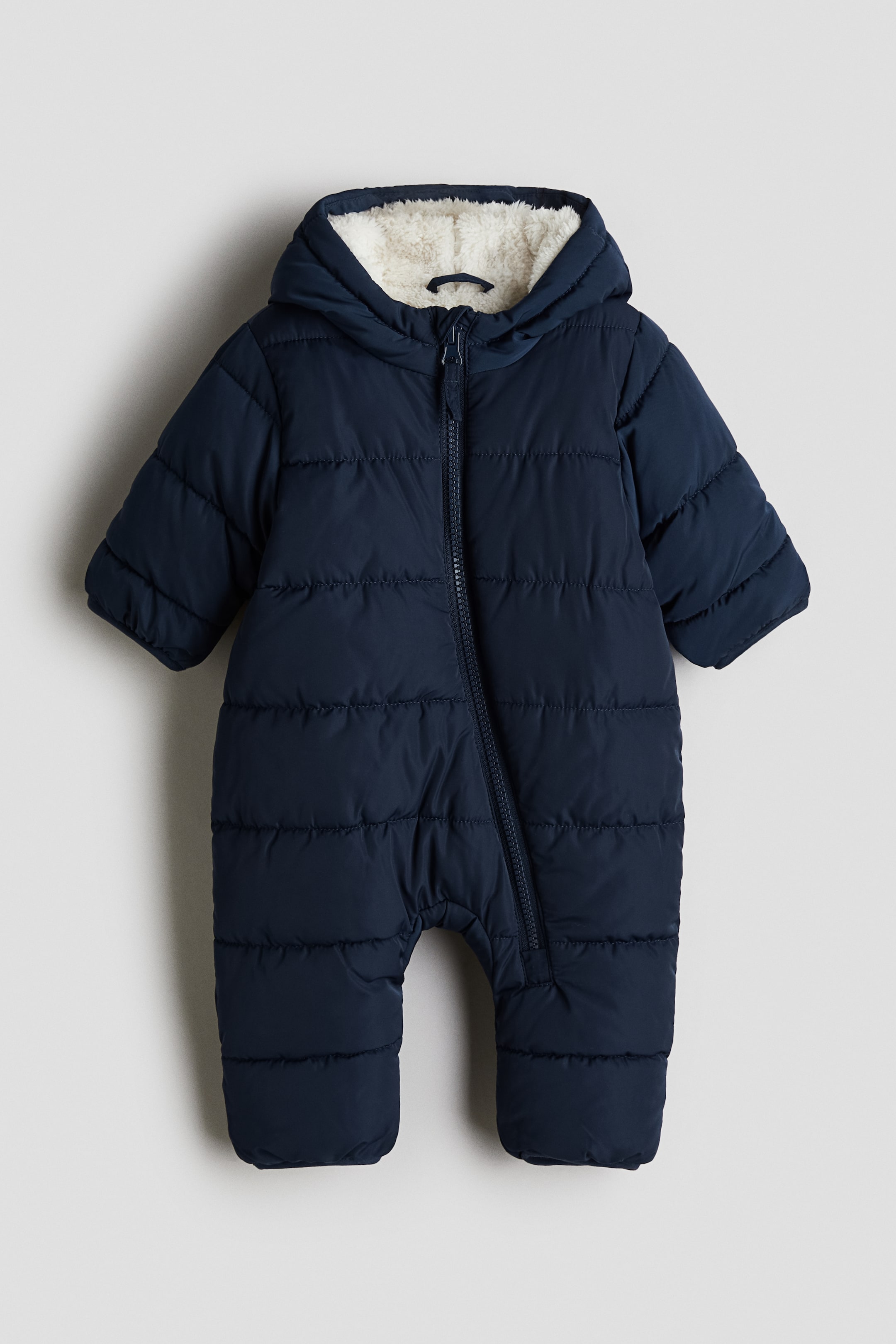 View larger image: A dark blue pile-lined snowsuit is displaying its FRONT, featuring horizontal quilting across its padded body and an attached hood with a visible cream-colored pile lining. A full-length zipper is running down the front and along the left leg, and the snowsuit is designed with open sleeve cuffs and integrated enclosed feet.