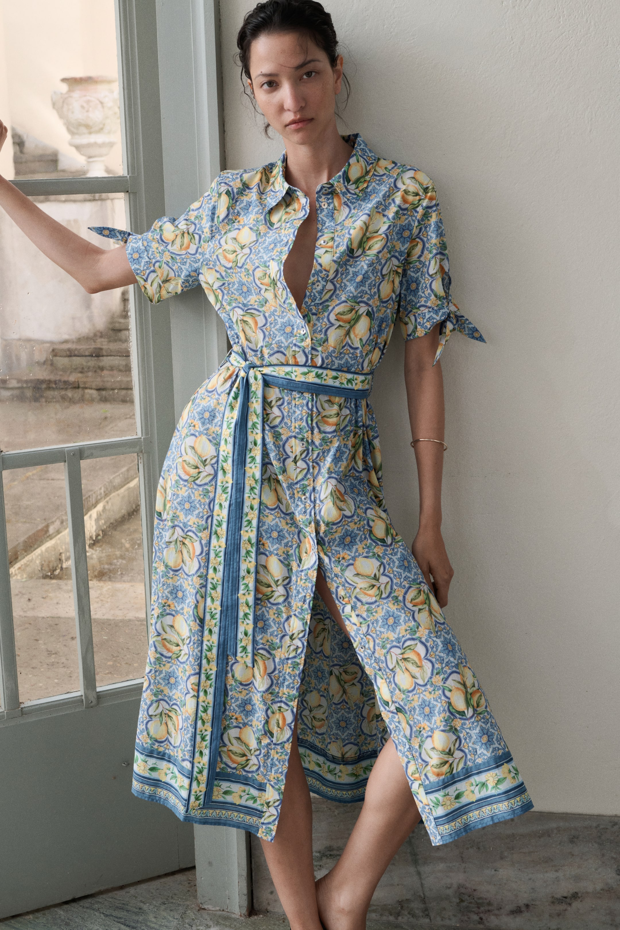 Größeres Bild ansehen: Das Midi-Hemdkleid, aus einem leicht front-rechten Winkel betrachtet, zeigt einen blauen und gelben Zitronen- und Blumenprint. Es verfügt über einen Hemdkragen und kurze Ärmel mit Bindedetails an den Manschetten. Das Design umfasst eine teilweise geöffnete Knopfleiste, einen passenden Stoffgürtel mit blau-gelbem Musterbesatz, der an der Taille gebunden wird, und einen Midirock mit einem hohen Schlitz auf der linken Seite und einem gemusterten Saumabschluss.