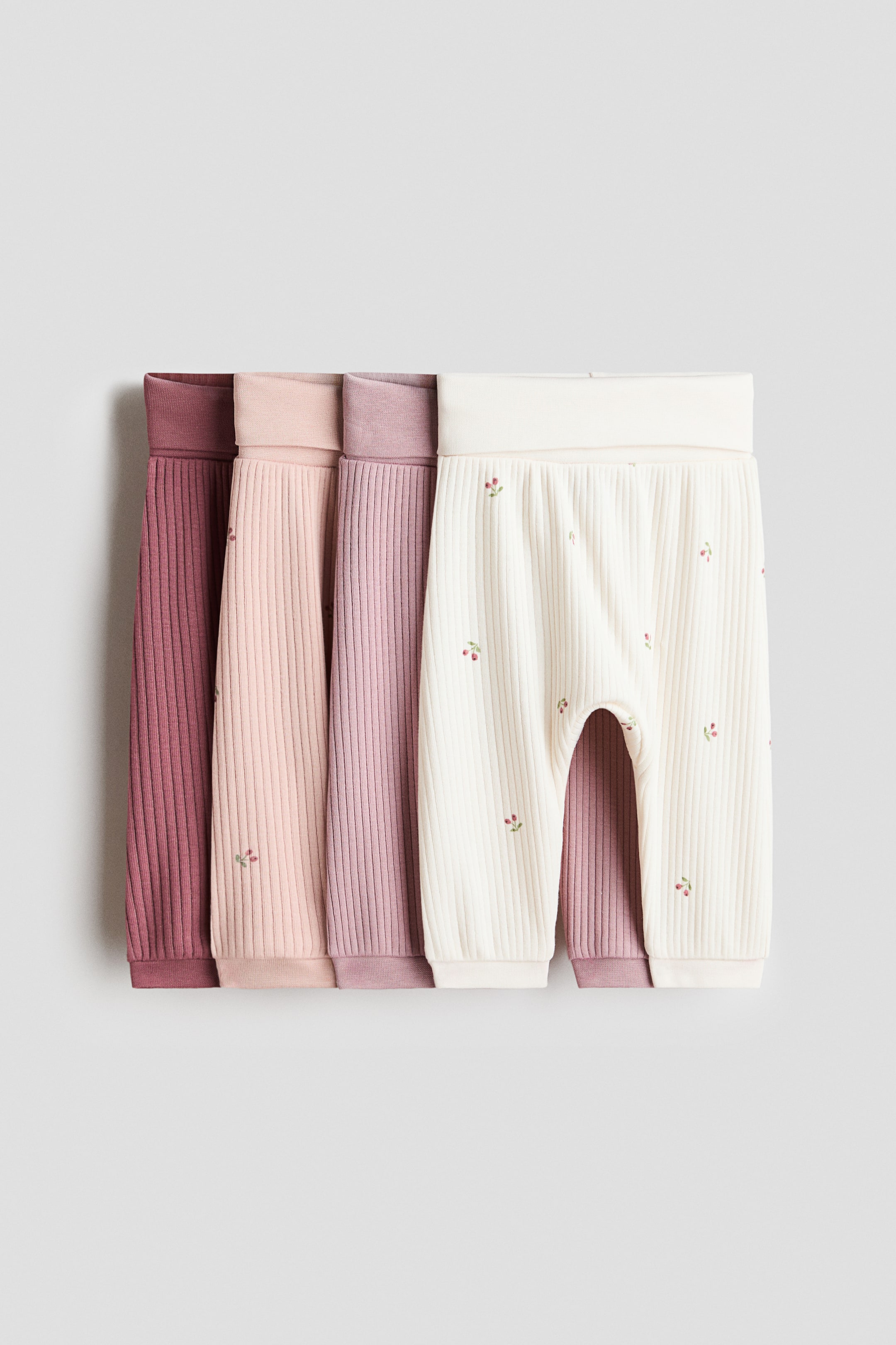 View larger image: Four pairs of soft, ribbed cotton jersey pants, each featuring a wide foldover panel at the waist, are standing upright. The frontmost cream pair is displaying its front, showing small embroidered cherry motifs and ribbed ankle cuffs, while the other pairs are visible in shades of pink and mauve.