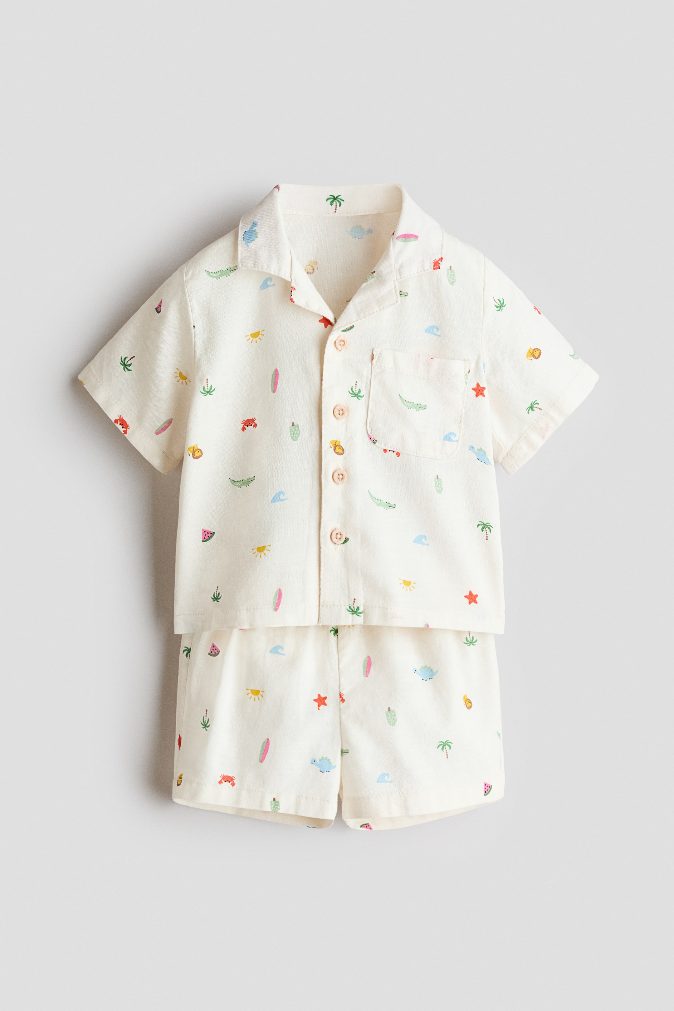 View larger image: A two-piece set, featuring a shirt and shorts, showcases a vibrant summer-themed print on off-white woven fabric. The straight-cut shirt is styled with a resort collar, light pink buttons down the front, short sleeves, and an open chest pocket, while the matching shorts offer a relaxed fit with an elastic waistband.