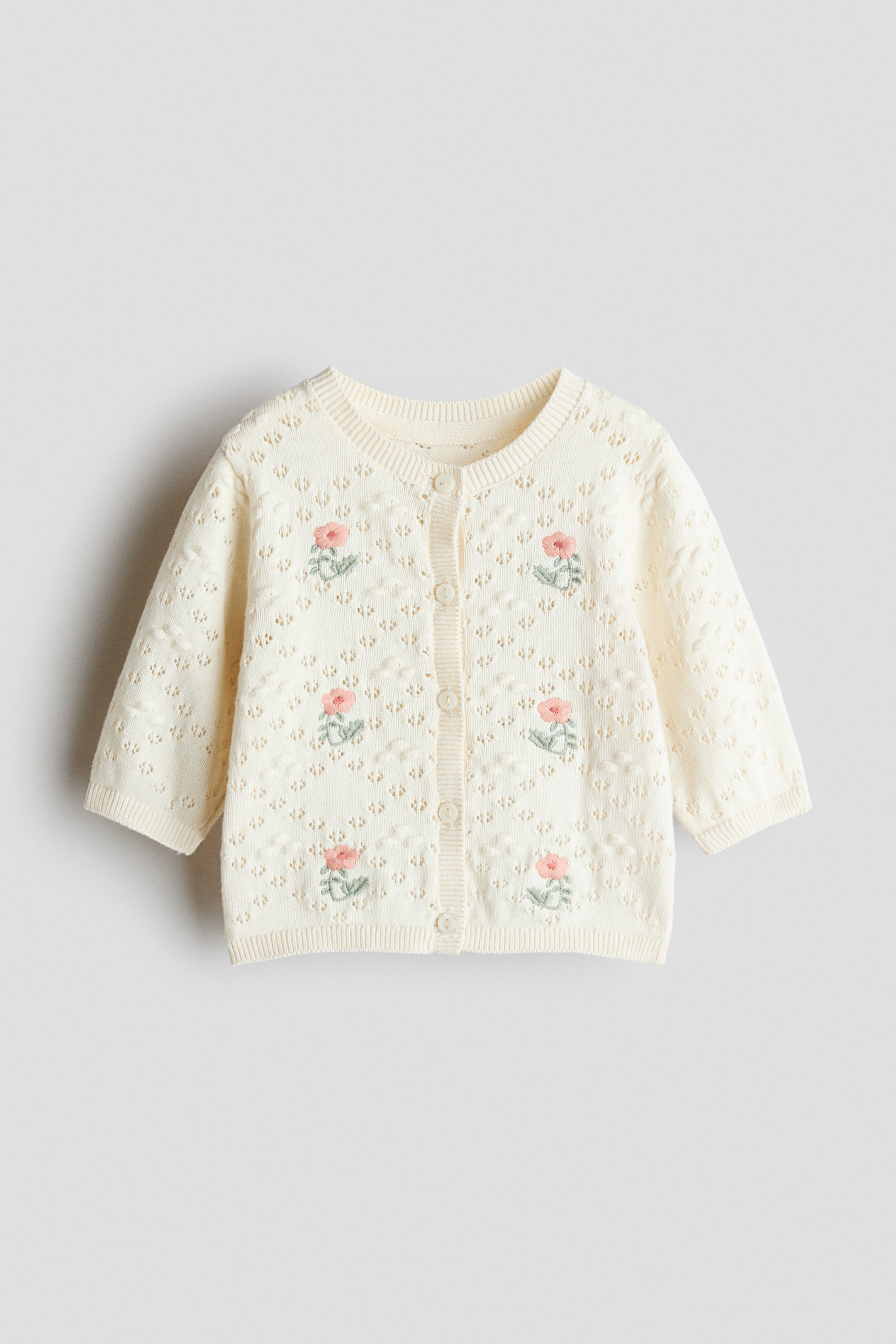 Visualizza immagine più grande: Un cardigan in maglia a punto traforato color crema è mostrato dal davanti, caratterizzato da piccoli fiori ricamati rosa, una chiusura a bottoni e bordi a coste sullo scollo, sui polsini e sull'orlo.
