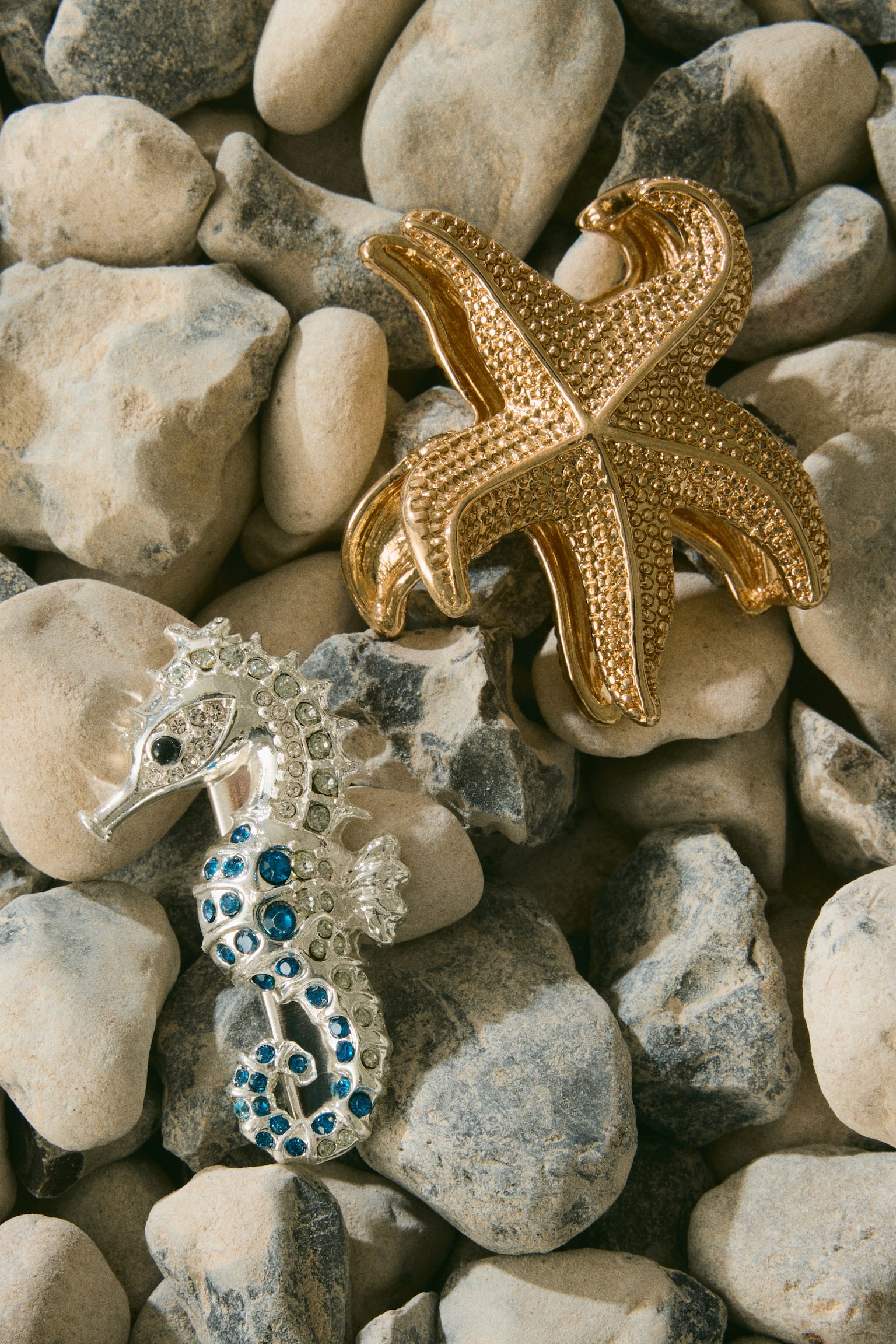 A golden starfish brooch is showcasing a textured surface with raised details on its five arms. A silver seahorse brooch is lying beside it, featuring clear and blue gemstones adorning its body and a black gemstone for its eye. - 1