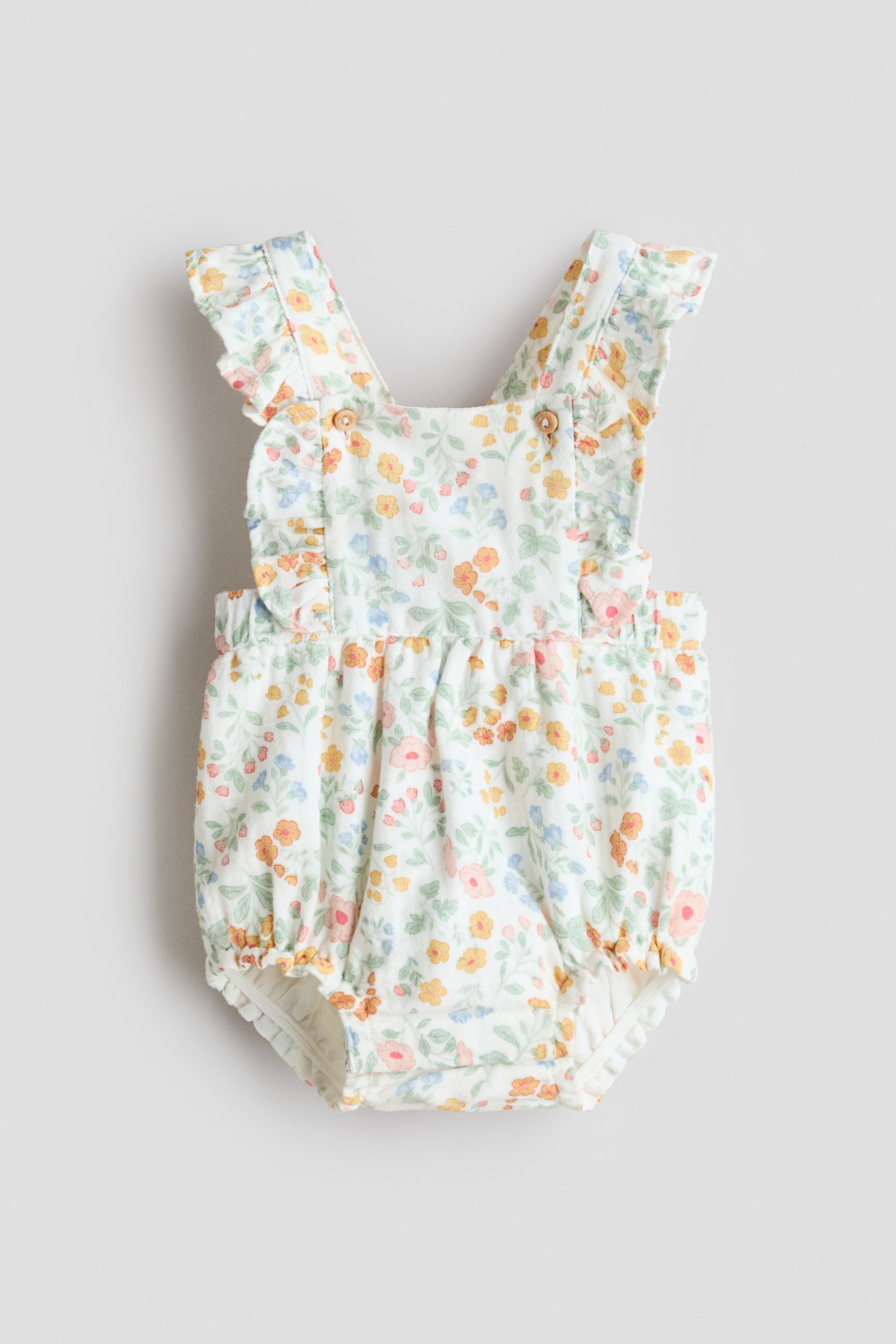 View larger image: A pair of overall shorts in a white woven cotton with a colorful floral pattern is facing FRONT. It features ruffle-trimmed shoulder straps with buttons at the top, an elastic waist, and elastic hems.