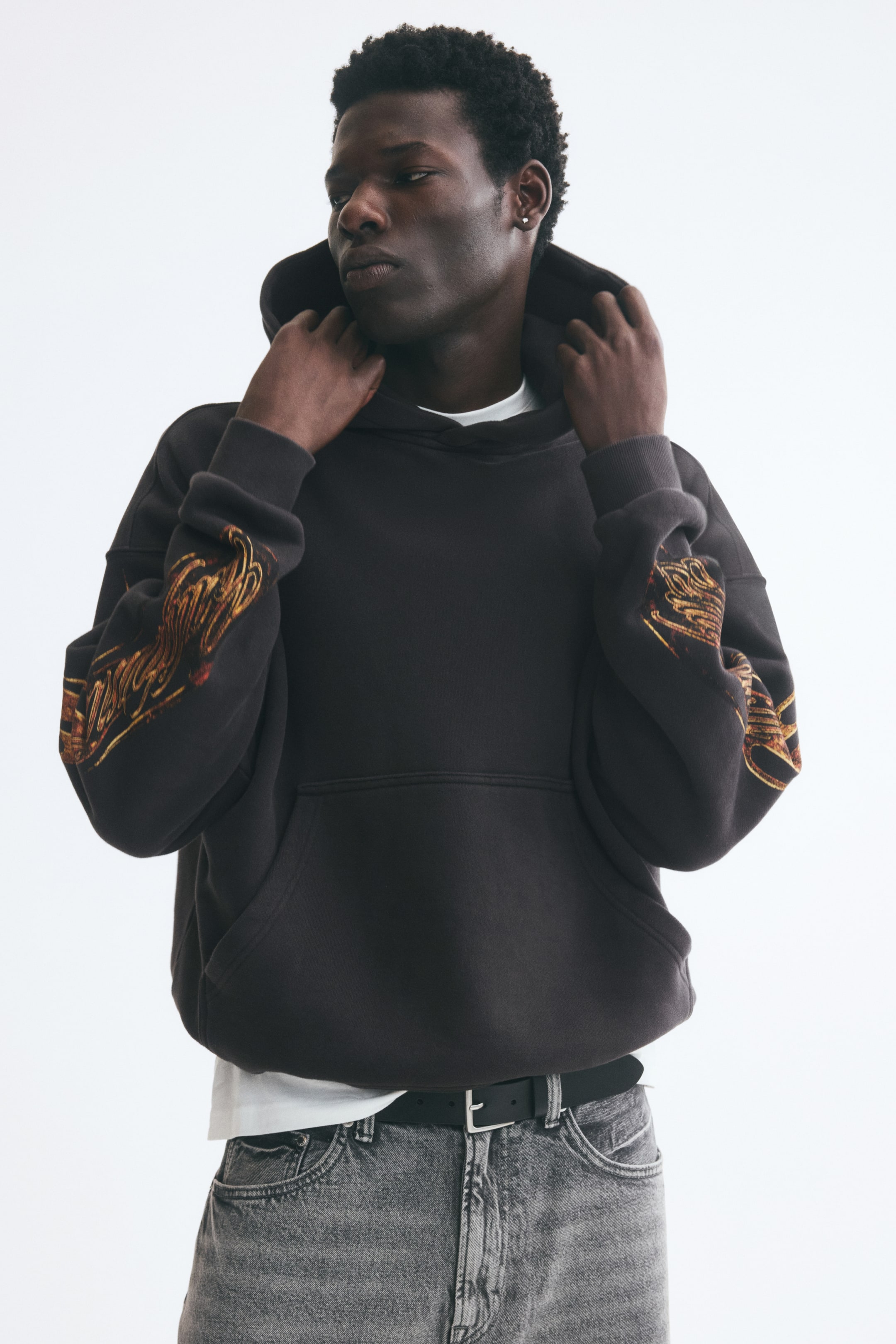 View larger image: A dark brown cotton hoodie with orange and yellow graphic prints on the sleeves is worn by the model, who is adjusting the wrap hood with both hands. The oversized hoodie features dropped shoulders, a kangaroo pocket, and ribbed cuffs, and is layered over a white T-shirt and gray jeans.