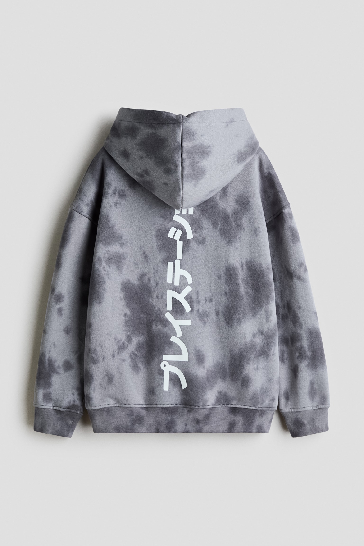 A loose-fit hoodie with a grey and dark grey tie-dye pattern is shown from the back, featuring a lined hood, dropped shoulders, and ribbed cuffs and hem. A vertical print of white Japanese characters runs down the centre of the back.