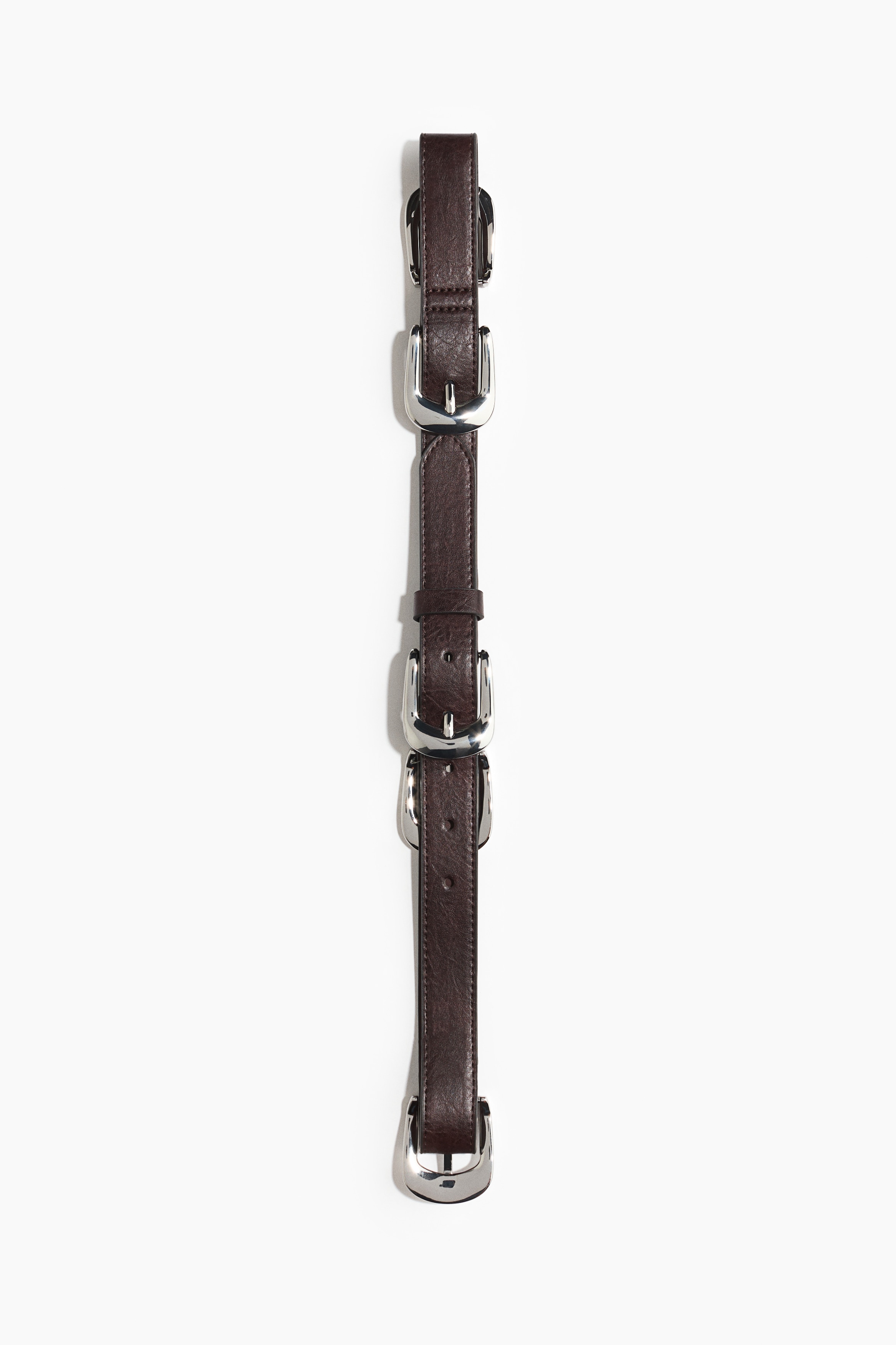 A dark brown belt in coated fabric, featuring multiple evenly spaced silver-tone metal buckles, is being worn through the loops of dark trousers by the model. The belt is styled with a tucked-in striped shirt and an open, floral-patterned jacket.
