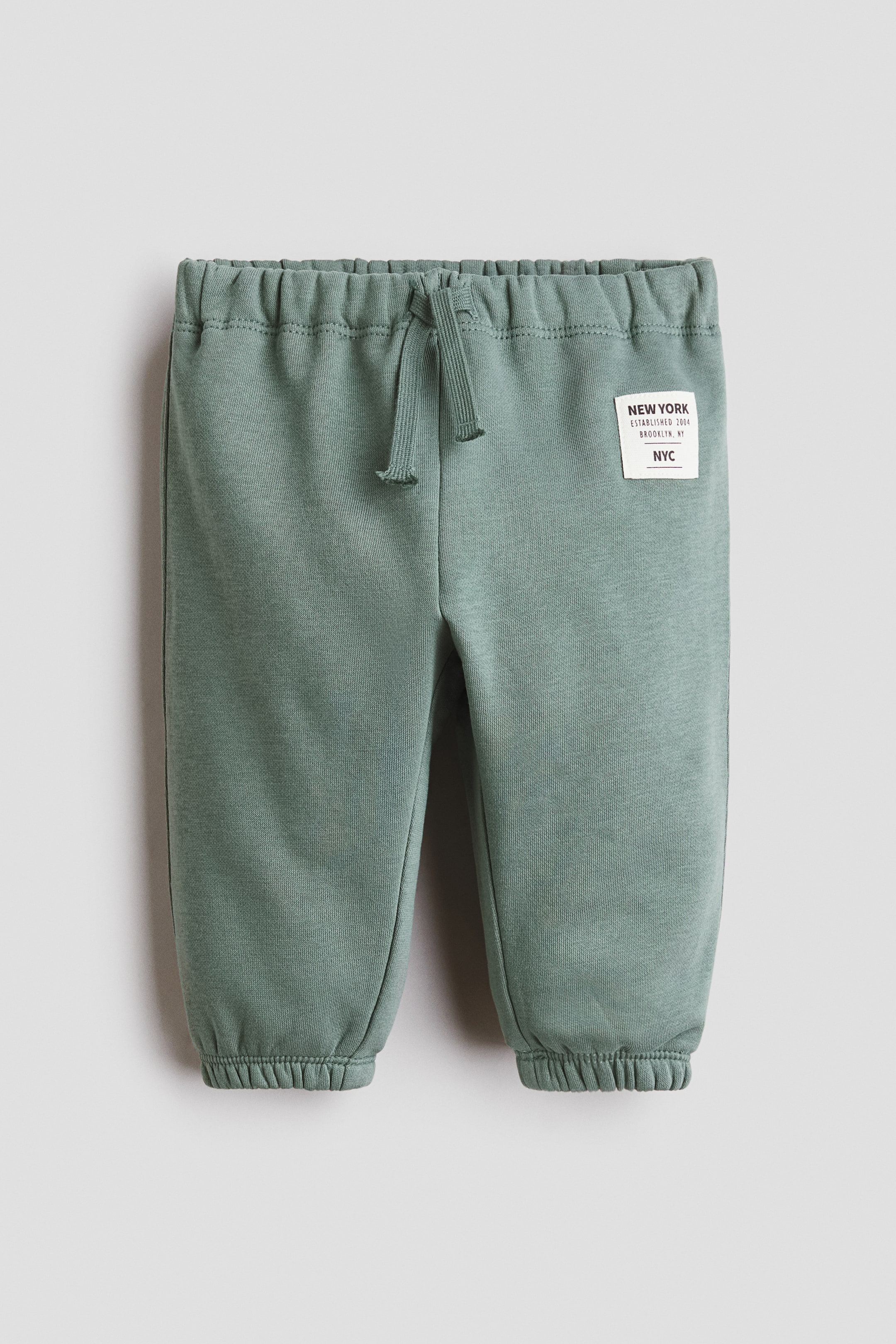 View larger image: "Muted green joggers in sweatshirt fabric are shown from the front, featuring an elasticated waist with a decorative drawstring. Covered elastication is visible at the hems, and a white rectangular label with text is on the upper right leg."