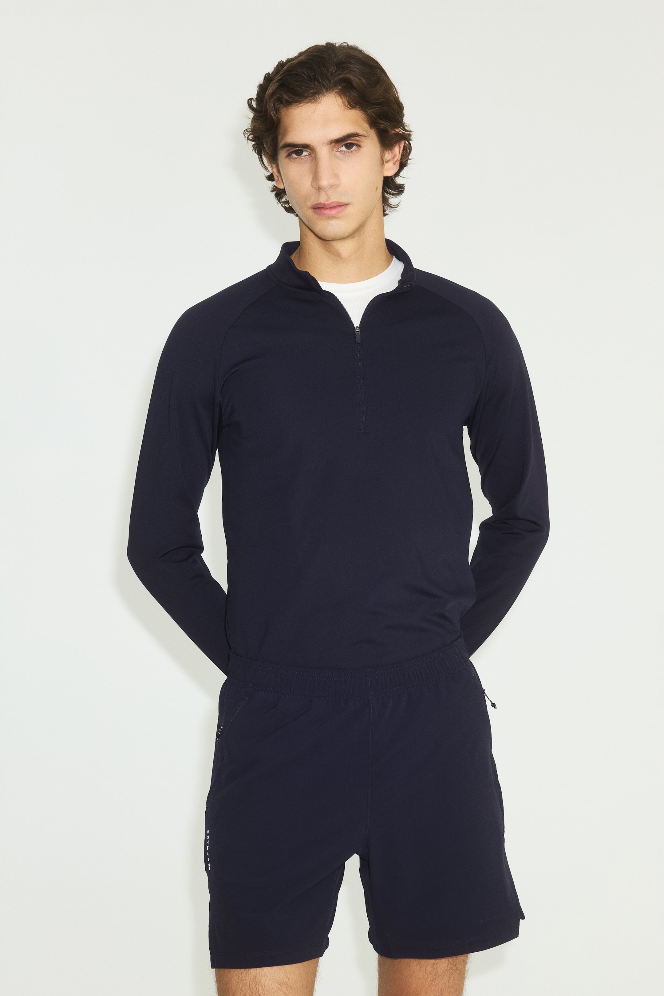 Se større billede: En marineblå halv-zip langærmet top har en stand-up krave, raglanærmer og en halv-zip lukning, med en hvid crew neck t-shirt synlig nedenunder. Den er parret med matchende marineblå shorts, der har en elastisk linning, sidelommer med lynlås og en diskret hvid grafik på venstre ben.