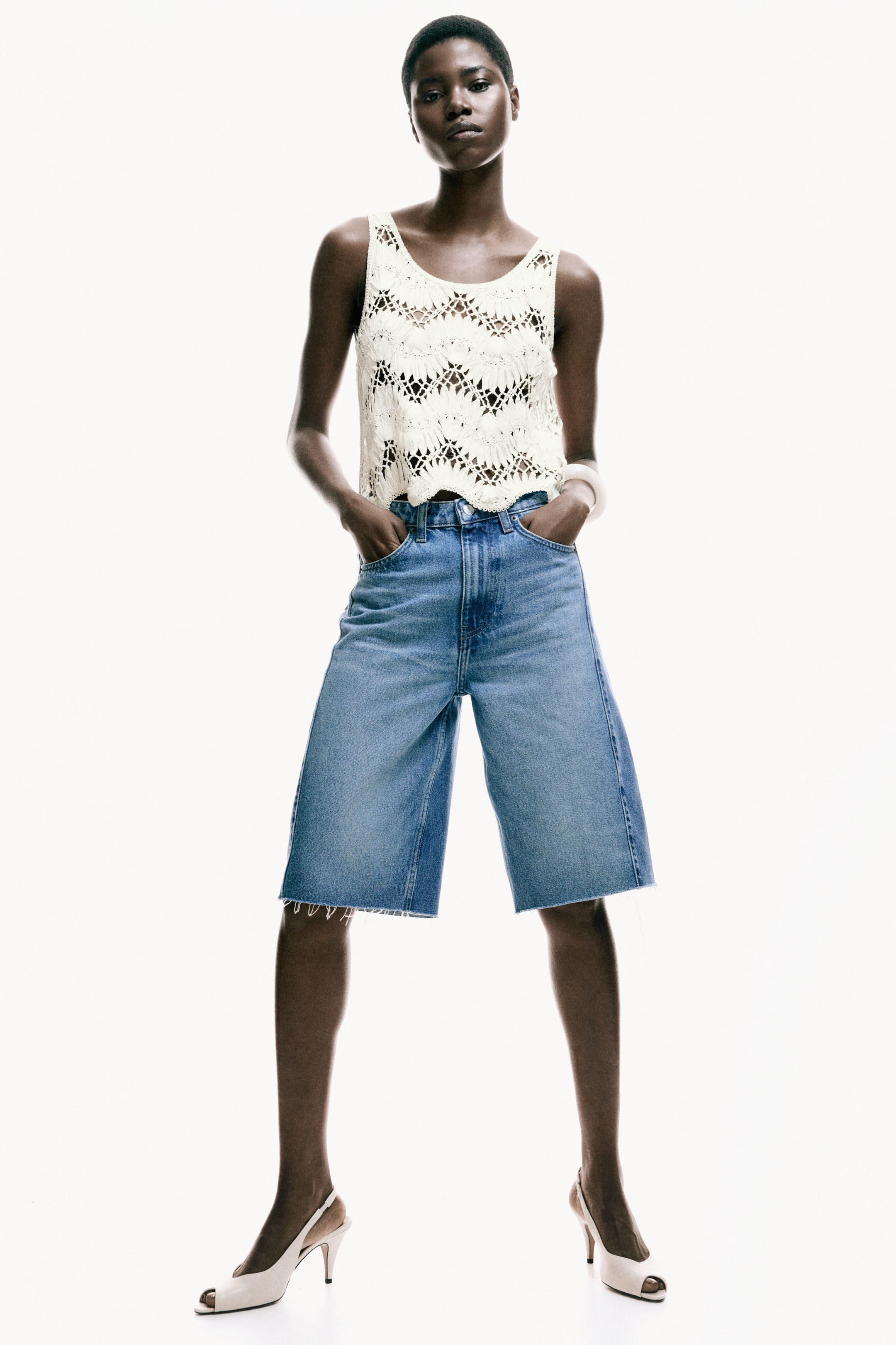 View larger image: A cream open-knit crochet tank top, featuring a geometric pattern and a scalloped hem, is styled with light wash, high-waisted denim Bermuda shorts showcasing a wide-leg fit and frayed raw hems. Cream slingback peep-toe high heels complete the ensemble.