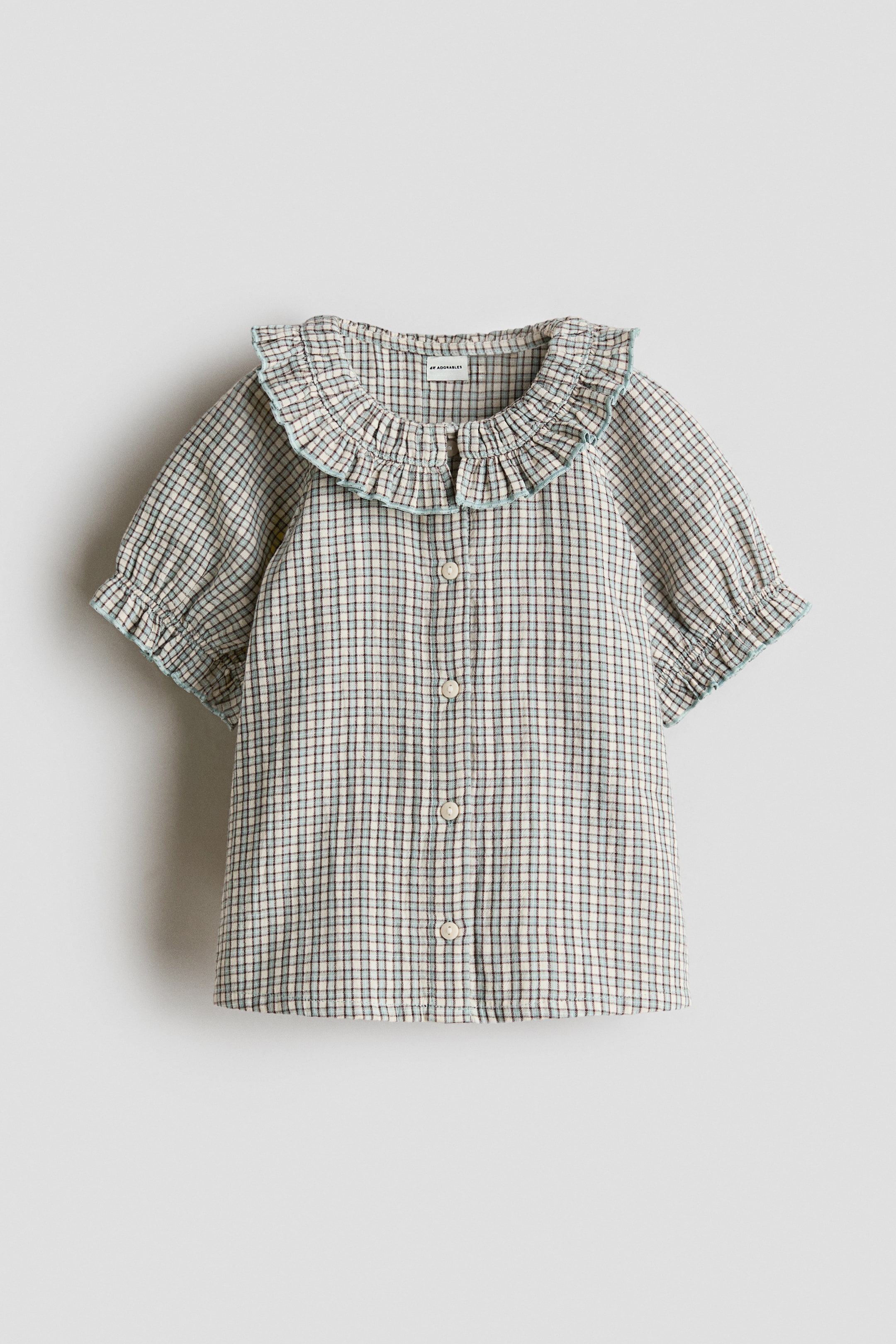 View larger image: A plaid shirt, facing front, showcases an off-white base adorned with blue and brown lines that create the pattern. It is crafted with a ruffled collar and short puffed sleeves, both highlighted with a light blue trim, and features a front button placket running down the centre.