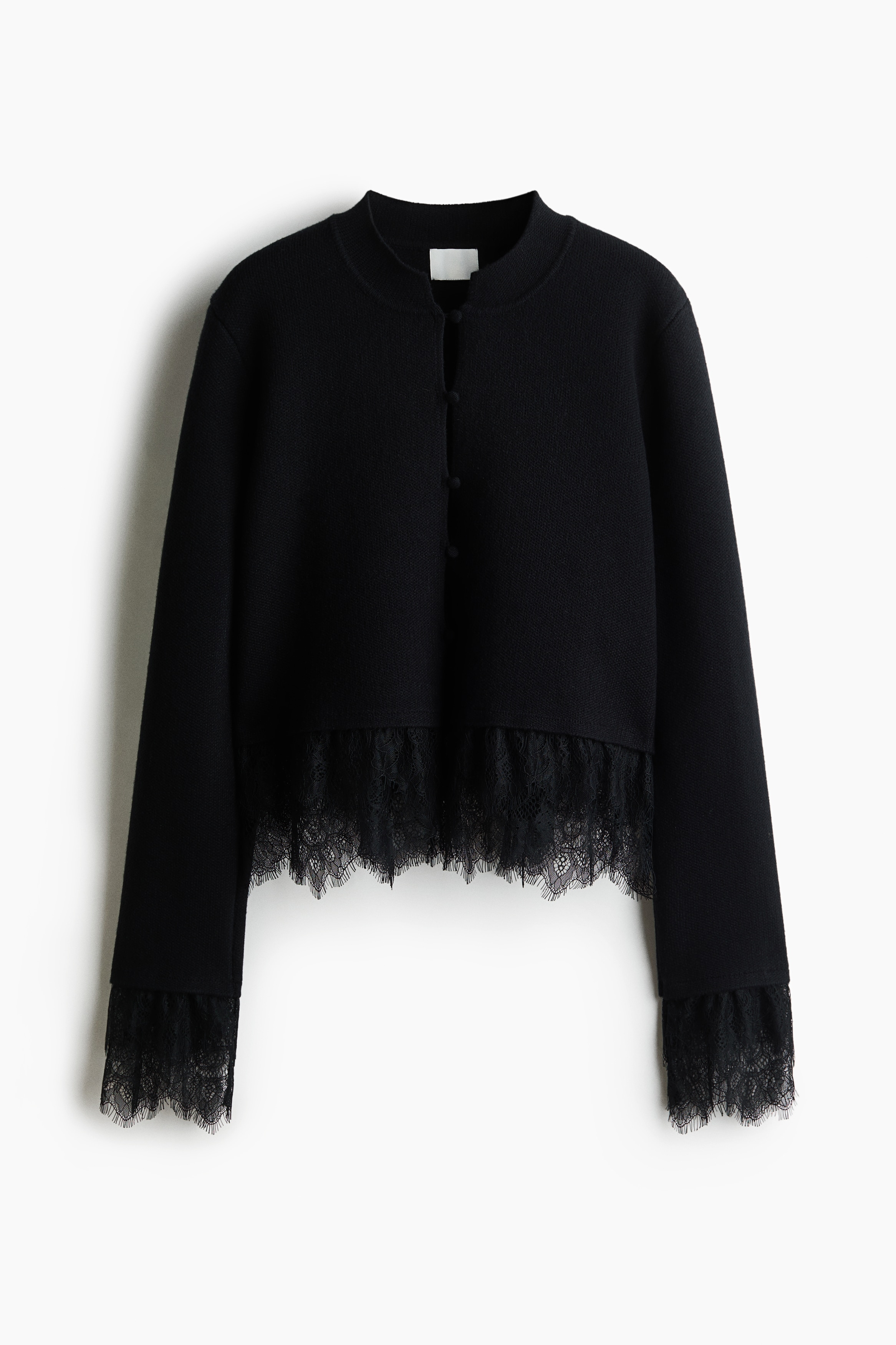 A black, short, fitted cardigan featuring a band collar, covered front buttons, and long sleeves is being worn by the model. The cardigan is detailed with black eyelash lace trim at the hem and cuffs, and is paired with matching black trousers and a belt with a large gold buckle.
