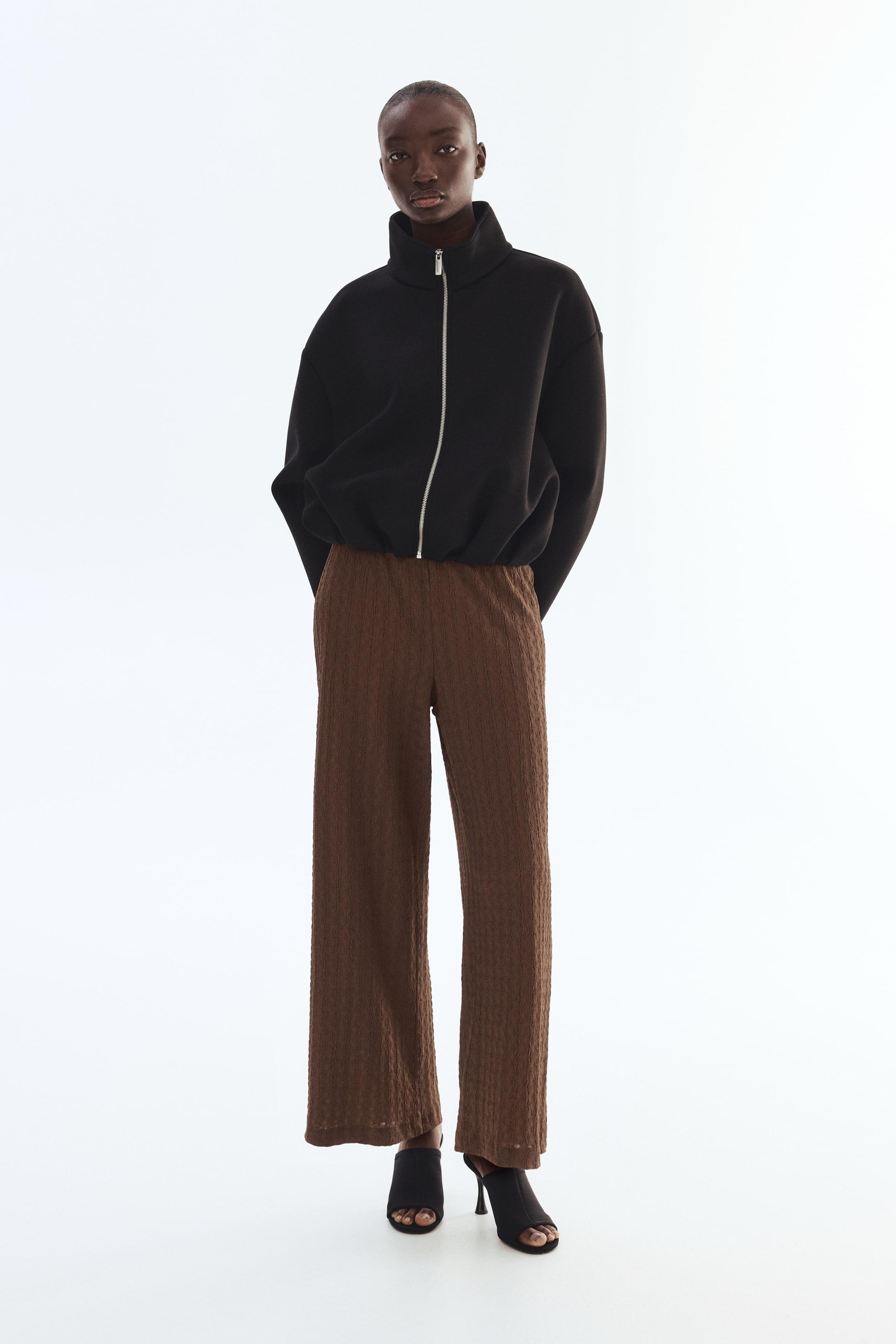 View larger image: A black high-neck jacket features a full-length silver zip and a cinched hem, offering a relaxed, blouson-style fit. Brown wide-leg trousers display a subtle textured pattern and an elasticated waist.