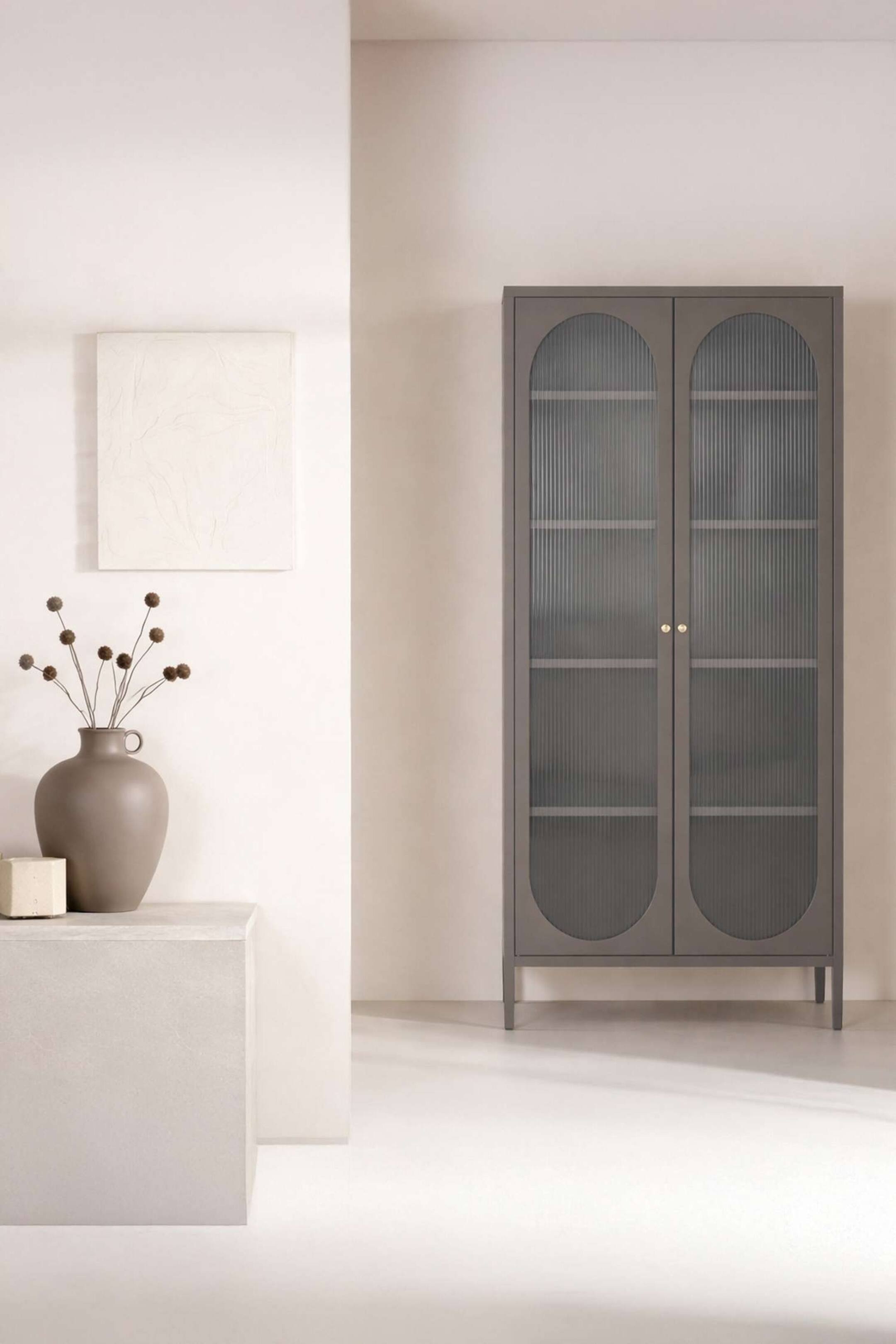 Agrandir l'image: L'armoire grise est dotée de deux portes, chacune avec un cadre supérieur arqué et un panneau inférieur semi-circulaire, encadrant un verre cannelé entre les deux. Elle est équipée de deux petites poignées rondes de couleur laiton et repose sur quatre pieds élancés.