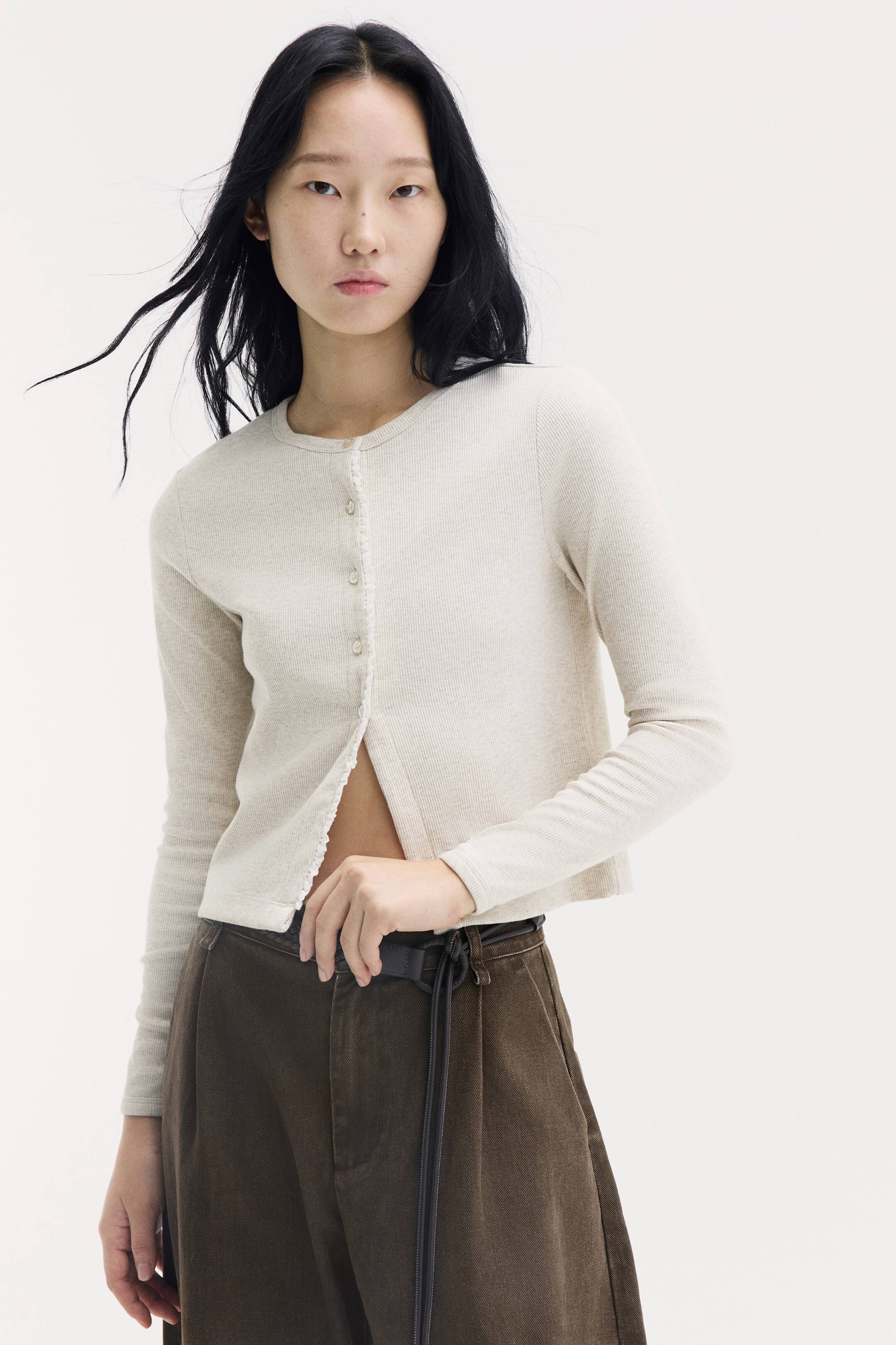 이미지 확대 보기: 모델이 연한 베이지색 리브드 저지 가디건을 착용하고 있으며, 라운드 넥라인과 긴소매, 그리고 버튼 플래킷을 따라 레이스 트리밍 디테일이 더해져 있습니다. 가디건은 상단만 버튼을 채운 상태로, 하단은 갈색 트라우저 위로 자연스럽게 오픈되어 있습니다.