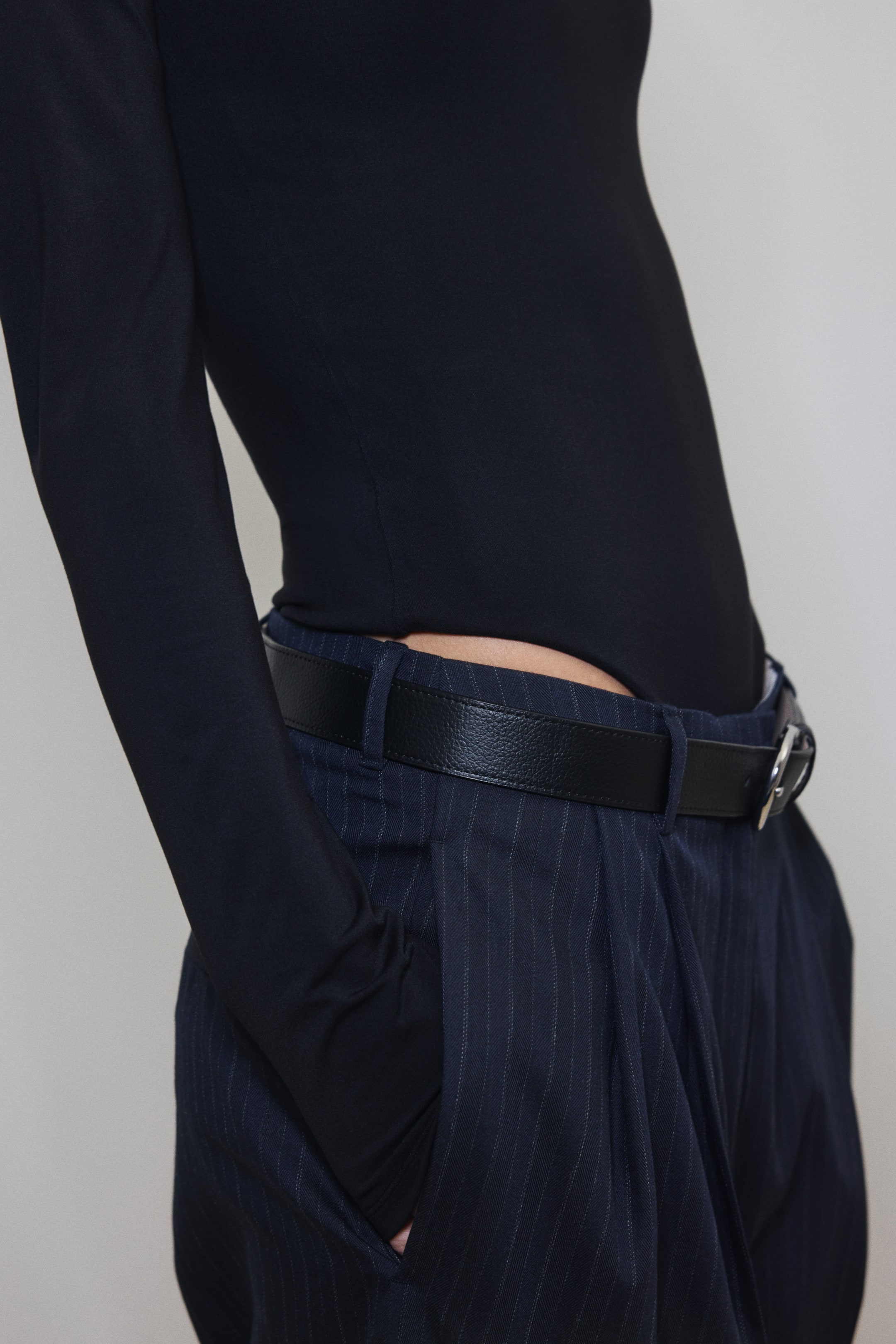 View larger image: A black, long-sleeved top is being worn, tucked into navy pinstripe trousers with pleats and a side pocket where a hand is resting. A black belt with a silver-toned buckle is cinching the trousers at the waist.
