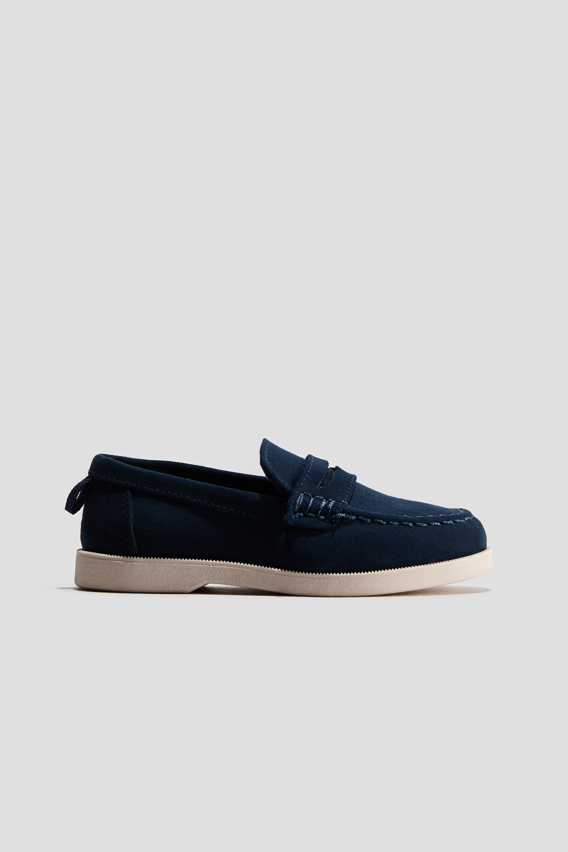 View larger image: The navy blue suede penny loafer is showcased in a side profile, highlighting a light beige sole with a textured edge and distinctive moccasin stitching encircling the toe box. A subtle fabric pull tab is also noticeable at the heel.
