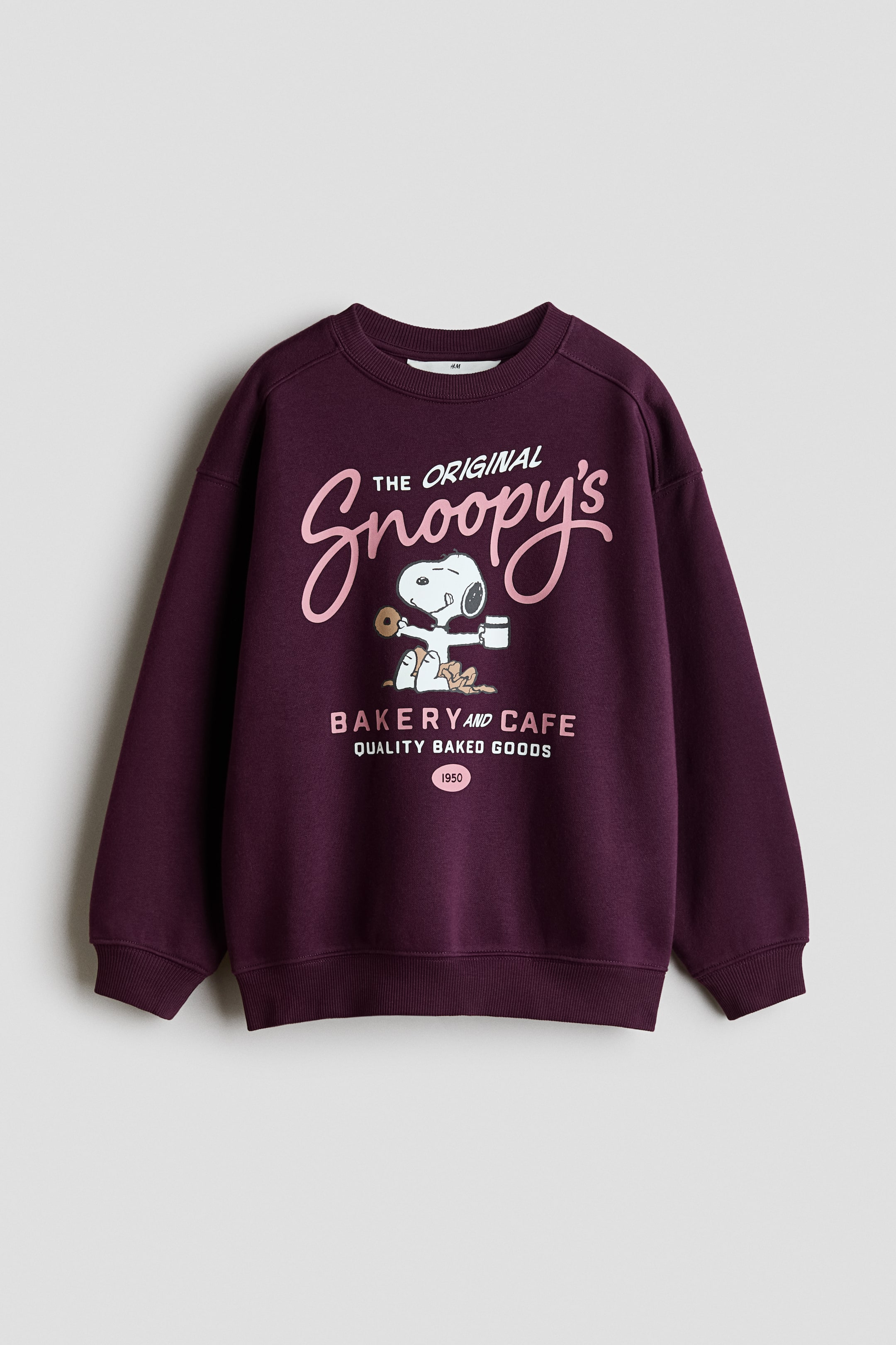 View larger image: A dark plum crew-neck sweatshirt is facing forward, featuring a graphic print on its front. The print is displaying "THE ORIGINAL Snoopy's" in pink script, with Snoopy holding a donut and coffee cup, above "BAKERY AND CAFE QUALITY BAKED GOODS 1950" in white and pink block letters; the sweatshirt is designed with long raglan sleeves, ribbed cuffs, and a ribbed hem.