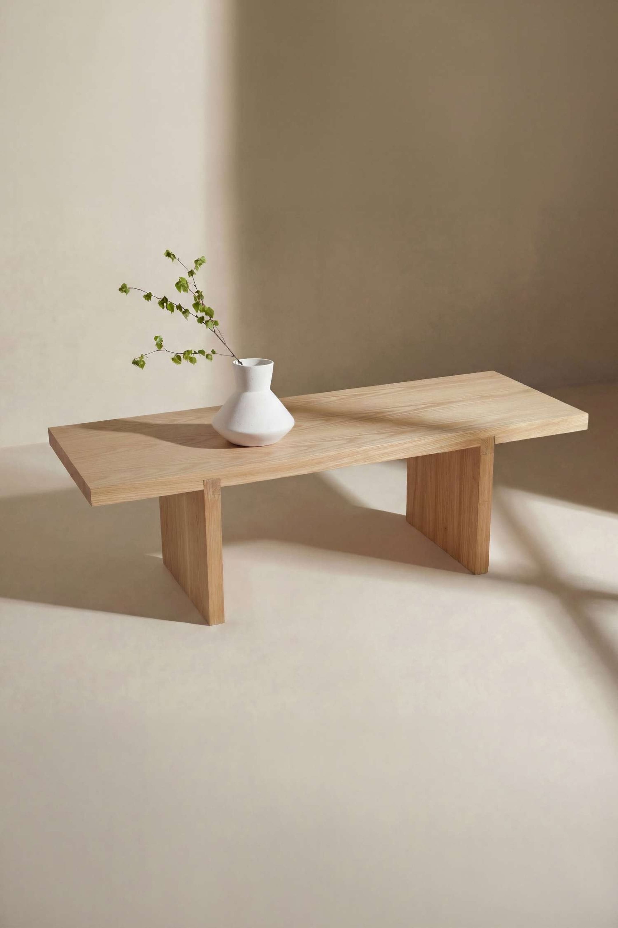 Agrandir l'image: Un banc en bois de couleur claire, présenté sous un angle trois-quarts, présente un plateau rectangulaire épais avec un grain visible, soutenu par deux pieds perpendiculaires de style monolithe. Un vase en céramique géométrique blanc, contenant une fine branche verte, repose sur la surface supérieure.