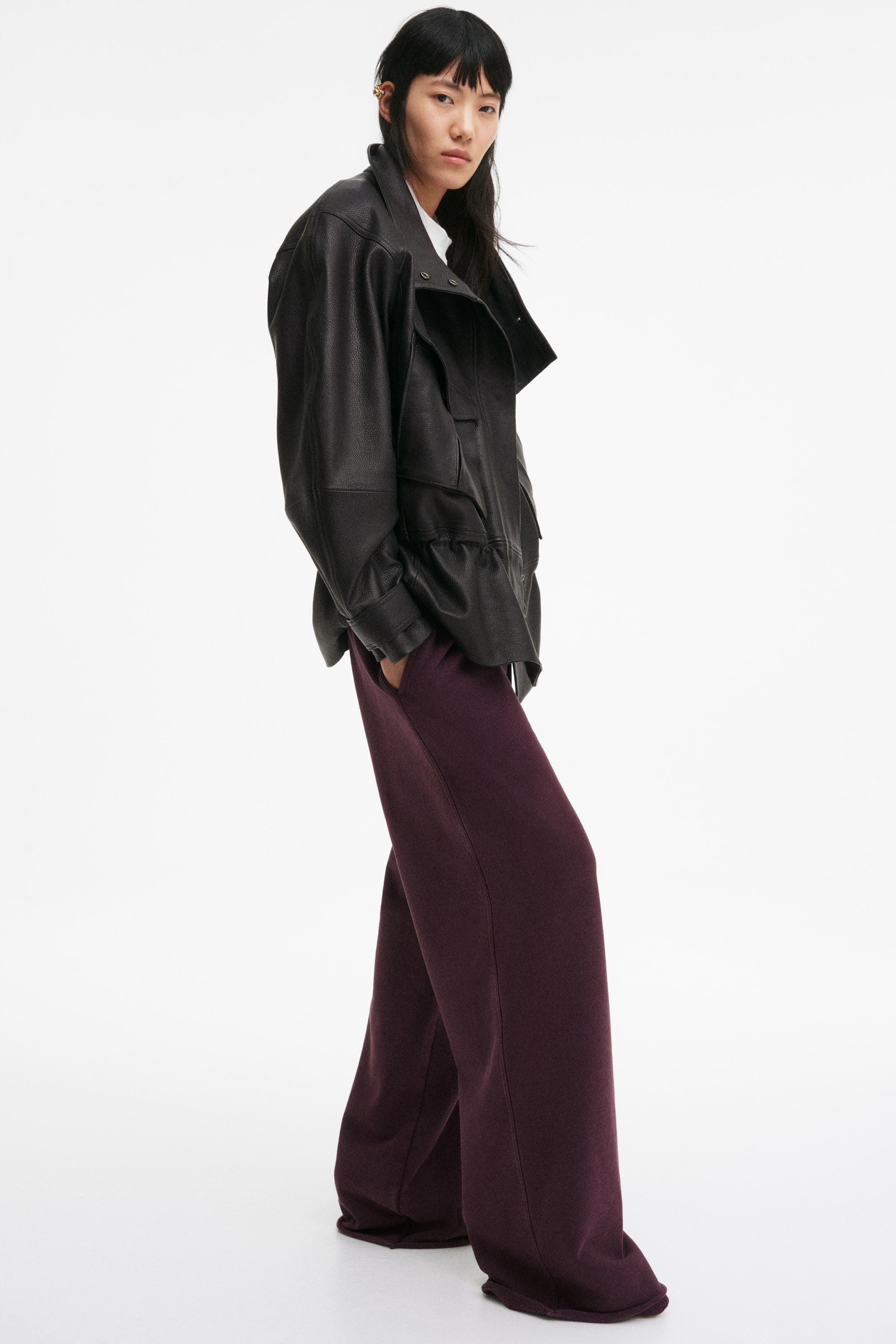 View larger image: A black pebbled leather jacket featuring a wide spread collar, a flap chest pocket, and an elasticated hem. Deep plum wide-leg trousers are worn, slightly pooling at the ankles.