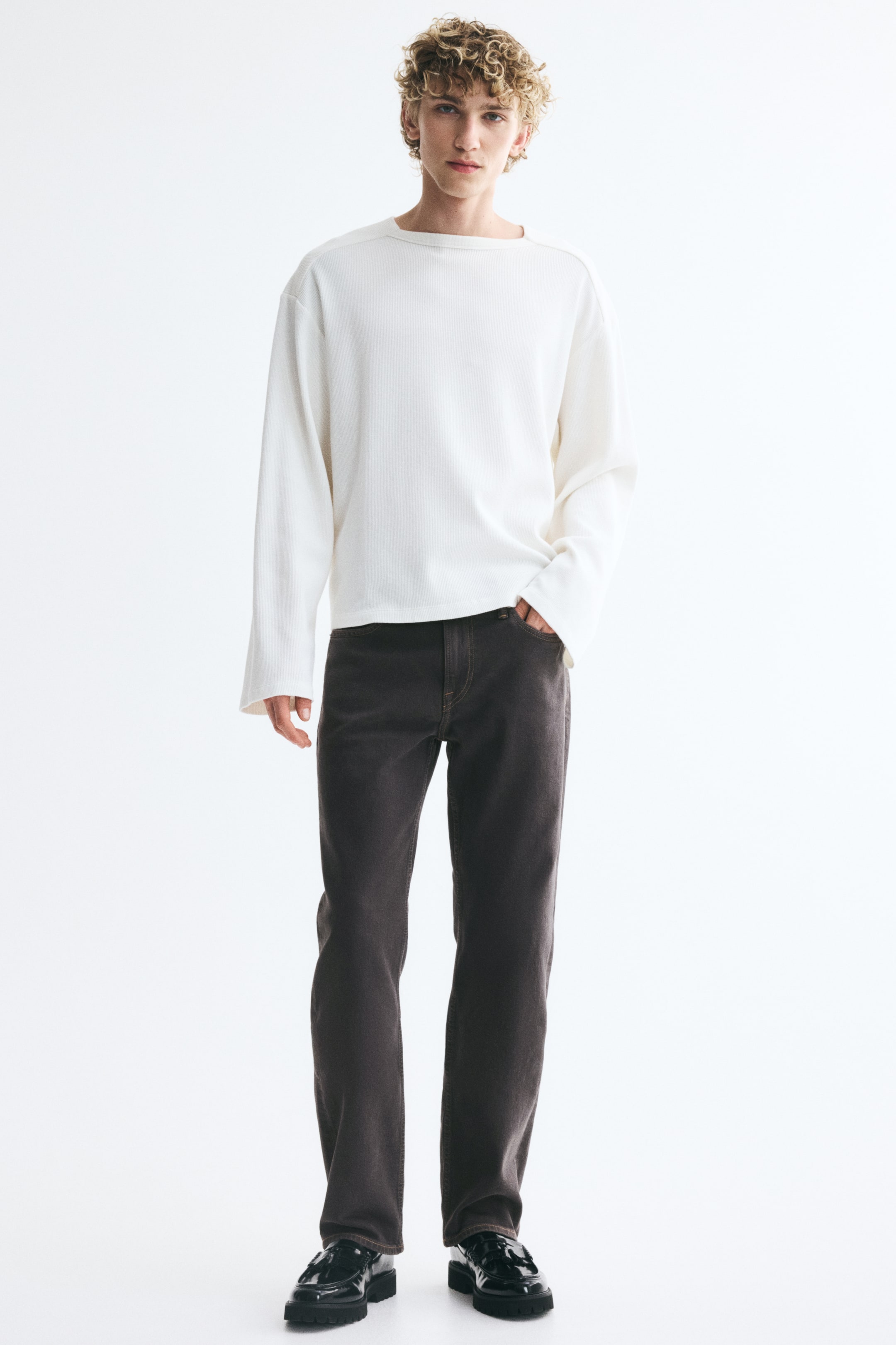 View larger image: Straight-leg jeans in dark brown denim are being worn by the model, who is standing with one hand resting in the front pocket. The regular-fit jeans are paired with a white long-sleeve top and black patent leather shoes.
