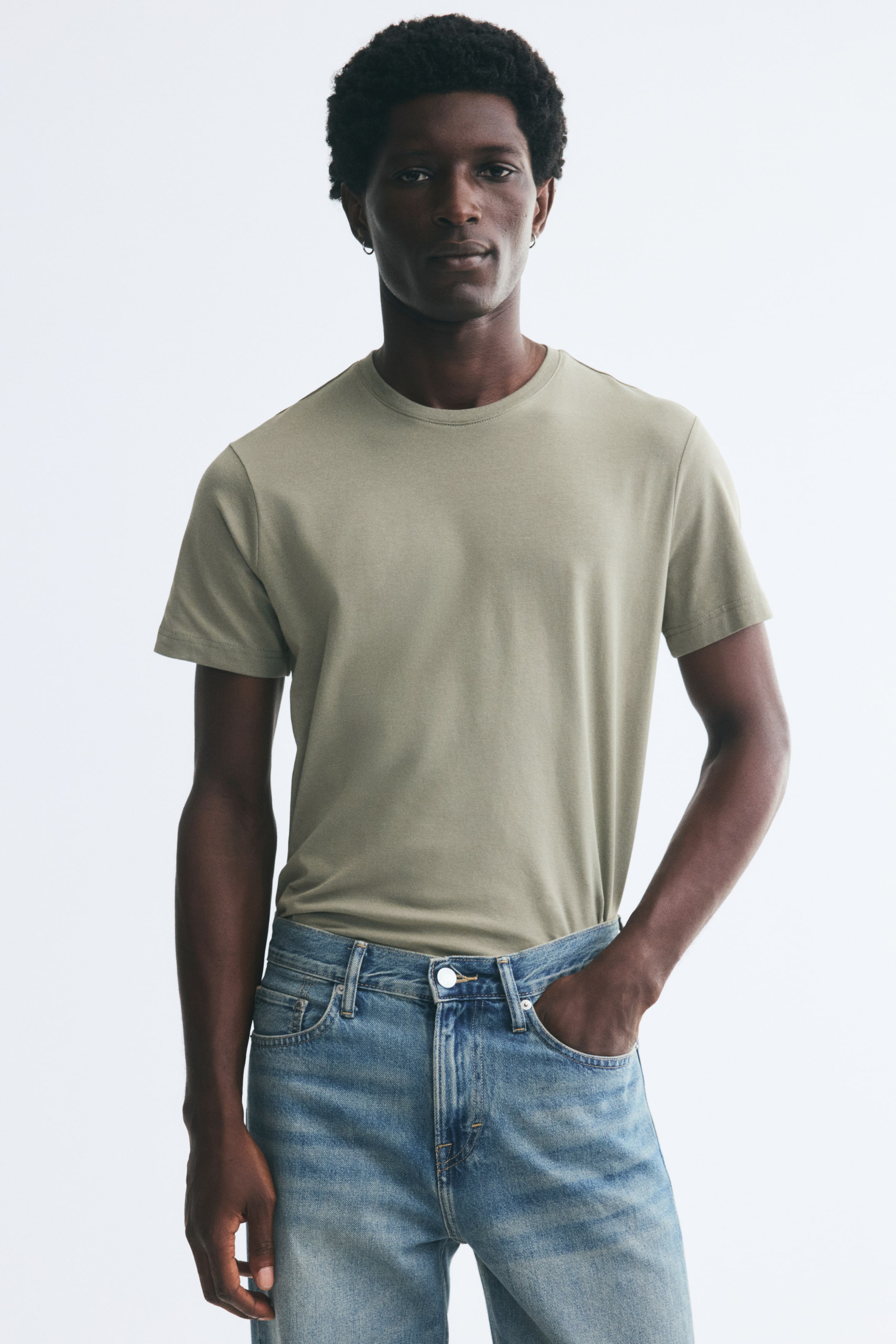 View larger image: The model is wearing a light sage green slim-fit T-shirt in lightweight cotton jersey, tucked into light-wash blue jeans. The tee features a round rib-trimmed neckline, short sleeves, and a fitted silhouette.