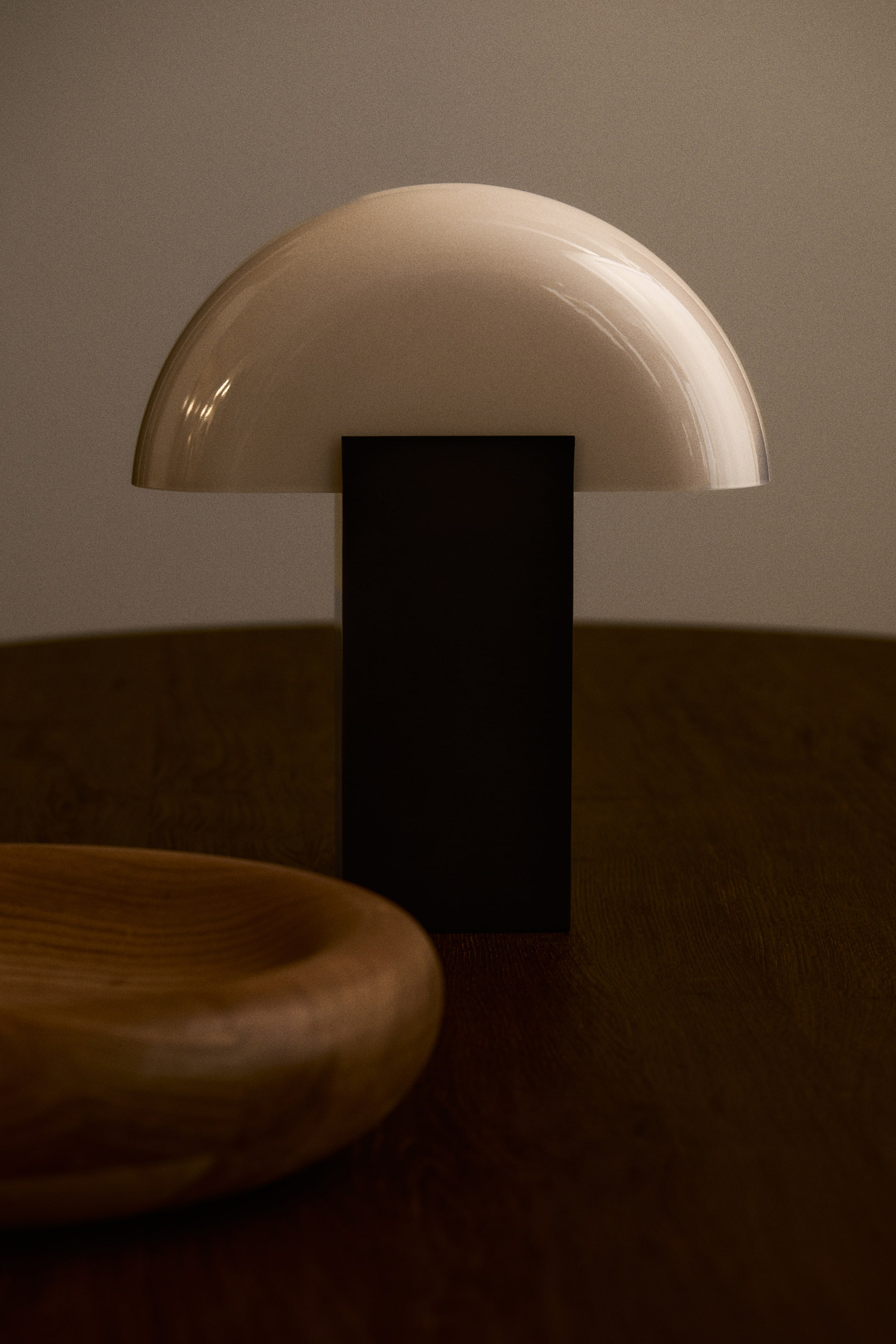 Ver imagen más grande: Una lámpara de mesa con un diseño geométrico se encuentra sobre una superficie de madera oscura, vista de frente. Presenta una pantalla de cristal en forma de cúpula, de color crema y acabado brillante, apoyada sobre una base rectangular de metal oscuro en forma de bloque.
