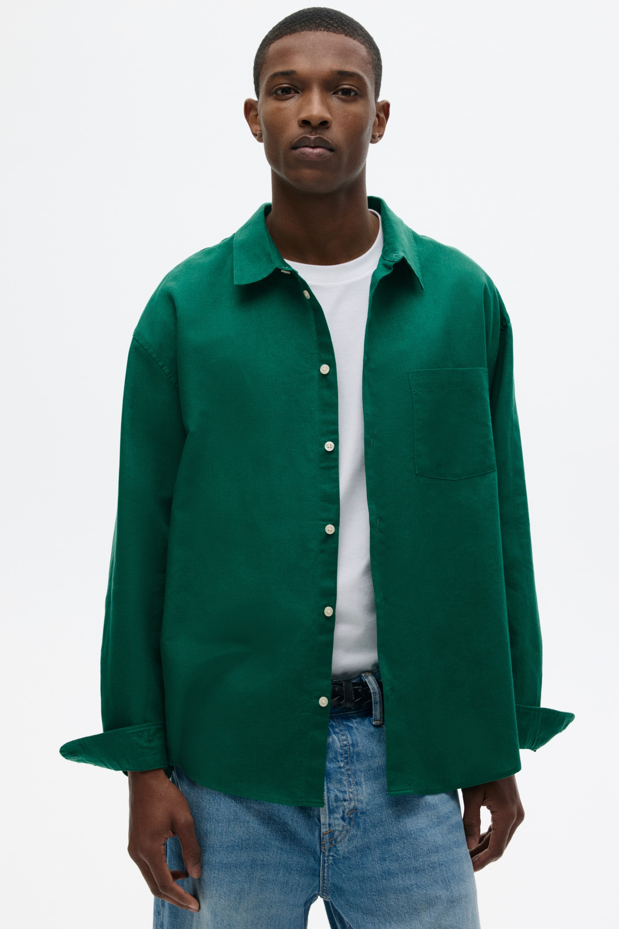 View larger image: A green button-front shirt is featuring a spread collar, a left chest patch pocket, and light-coloured buttons. The shirt is being worn open over a white T-shirt, with its long sleeves rolled up at the cuffs, and its hem styled untucked over light-wash jeans.