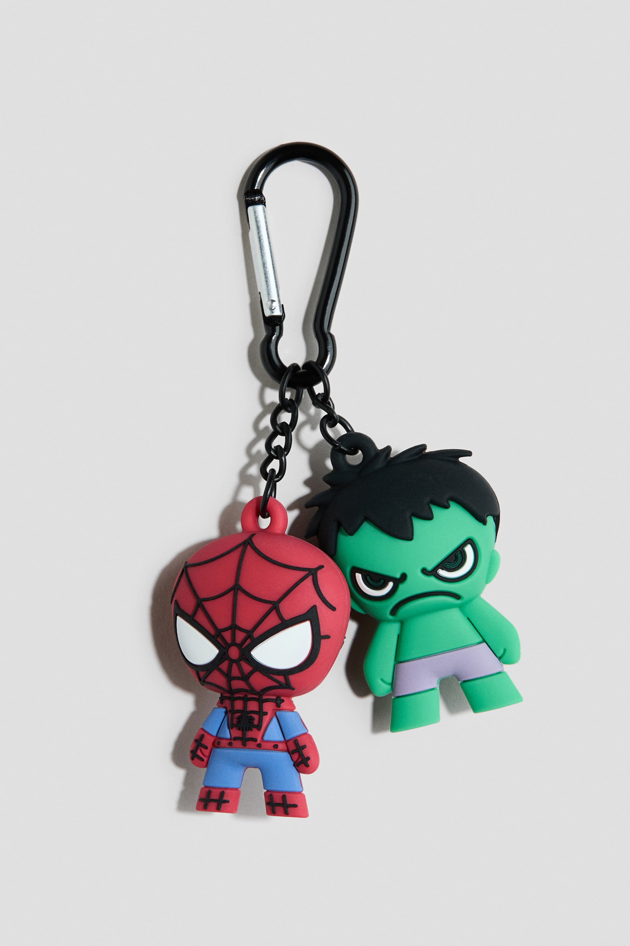 Ver imagem maior: Um pendente de mala apresenta um mosquetão preto do qual pendem duas figuras decorativas, ambas voltadas para a frente. A primeira figura representa o Homem-Aranha com uma cabeça vermelha, padrões de teia preta, olhos brancos e um corpo azul e vermelho, enquanto a segunda figura representa o Hulk com pele verde, cabelo preto, olhos brancos zangados e calções roxos.