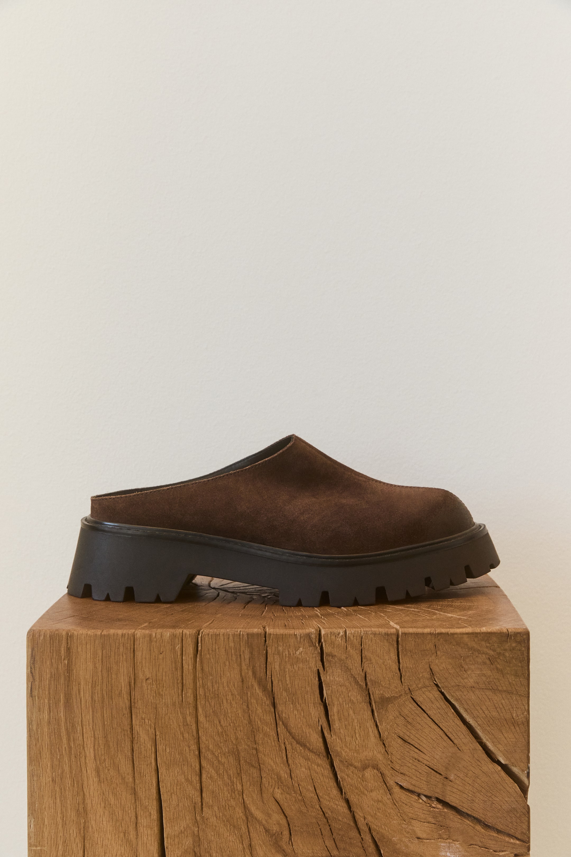 Ampliar la imagen: Un mule tipo loafer de gamuza café oscuro se muestra en vista de perfil lateral, resaltando una punta redondeada y un diseño slip-on de talón abierto. El zapato cuenta con una suela gruesa negra con dentado profundo y una banda de rodadura con patrón marcado.
