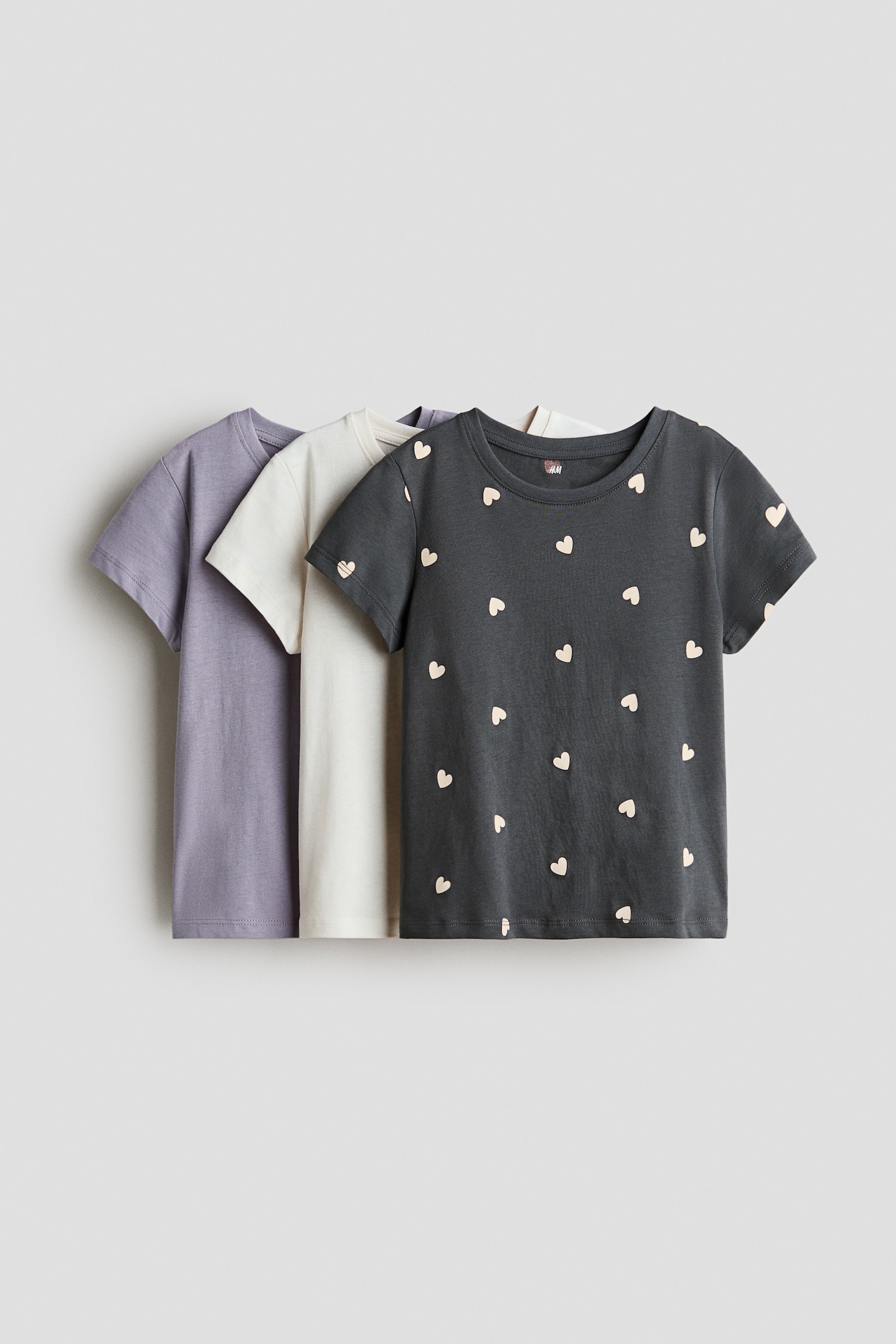 Three short-sleeved cotton jersey T-shirts are shown front-facing in a layered presentation. The front T-shirt is dark grey and features an all-over pattern of small light-coloured hearts, while the middle T-shirt is solid off-white and the back T-shirt is solid light purple, with all three featuring a round neckline with a narrow trim.