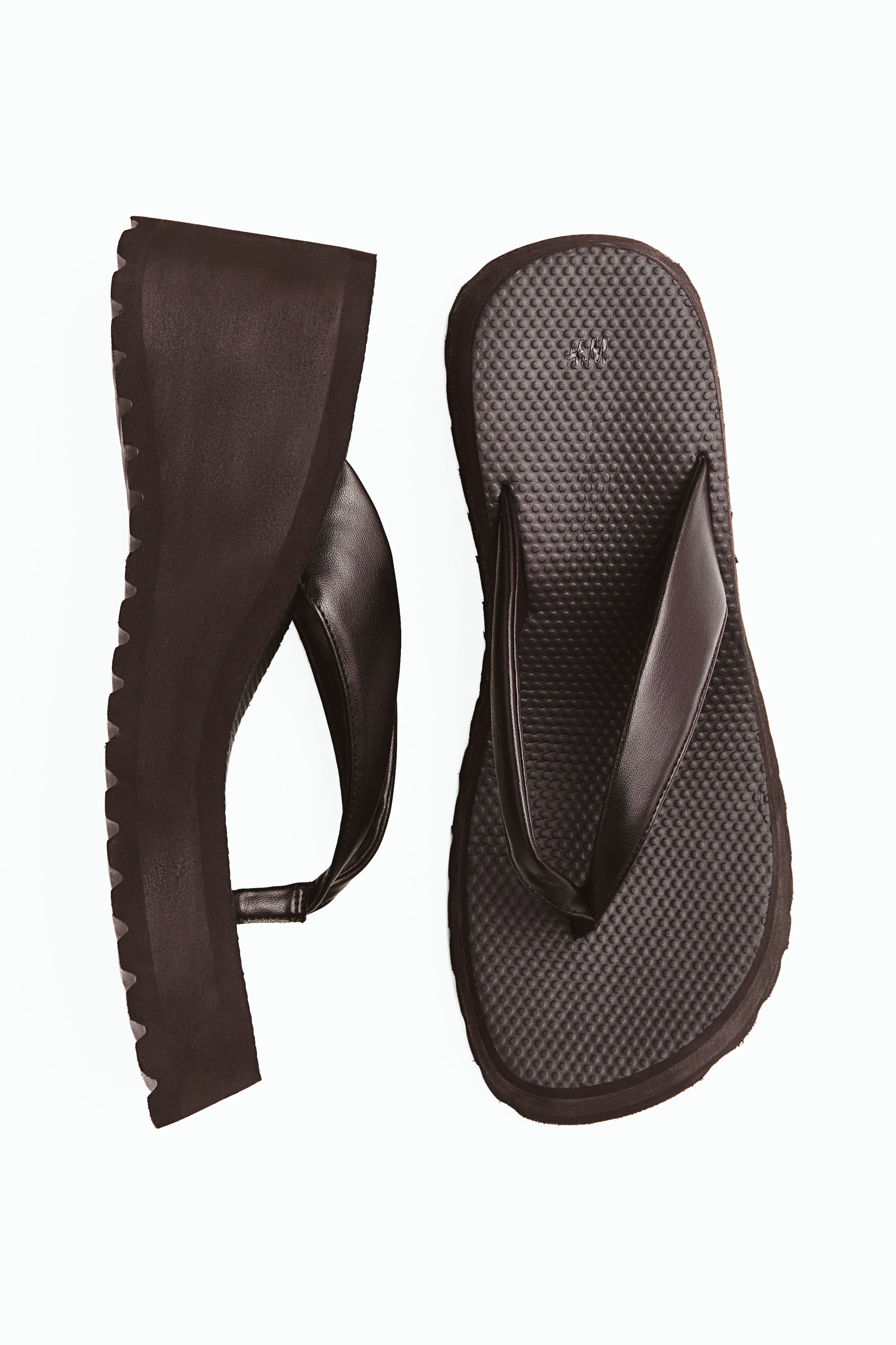 View larger image: Dark brown flip-flops are displayed, featuring wide foot straps, narrow toe posts, and oblique toes. One flip-flop lies flat, showcasing its textured footbed, while the other is positioned on its side, revealing its wedge heel with a ribbed pattern underneath.
