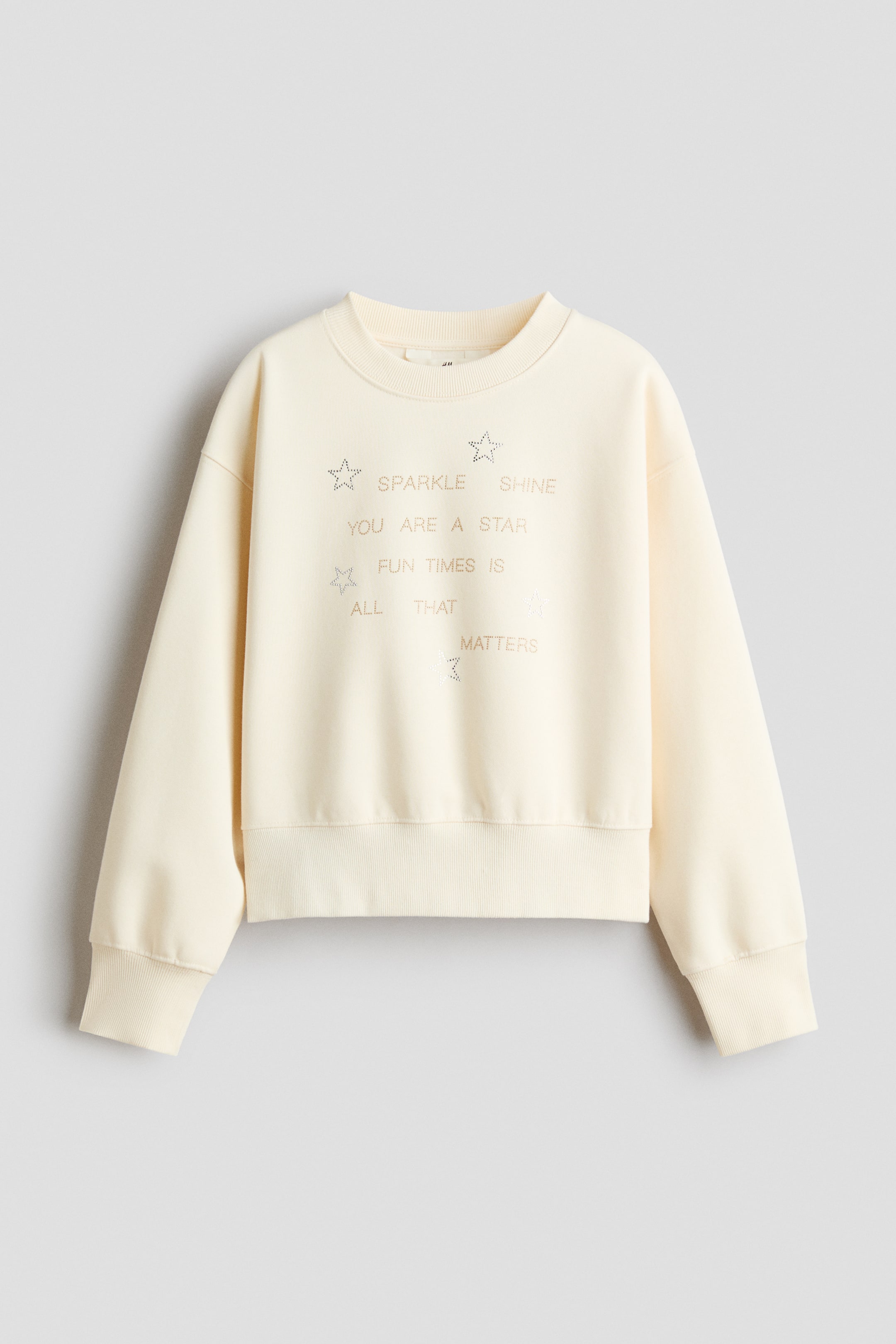 View larger image: A cream-colored, loose-fit fleece sweater is shown from the front, featuring a round rib-trimmed neckline, dropped shoulders, and wide ribbed cuffs and hem. The chest is adorned with shimmering rhinestone details forming star outlines and text that reads "SPARKLE SHINE," "YOU ARE A STAR," and "FUN TIMES IS ALL THAT MATTERS."