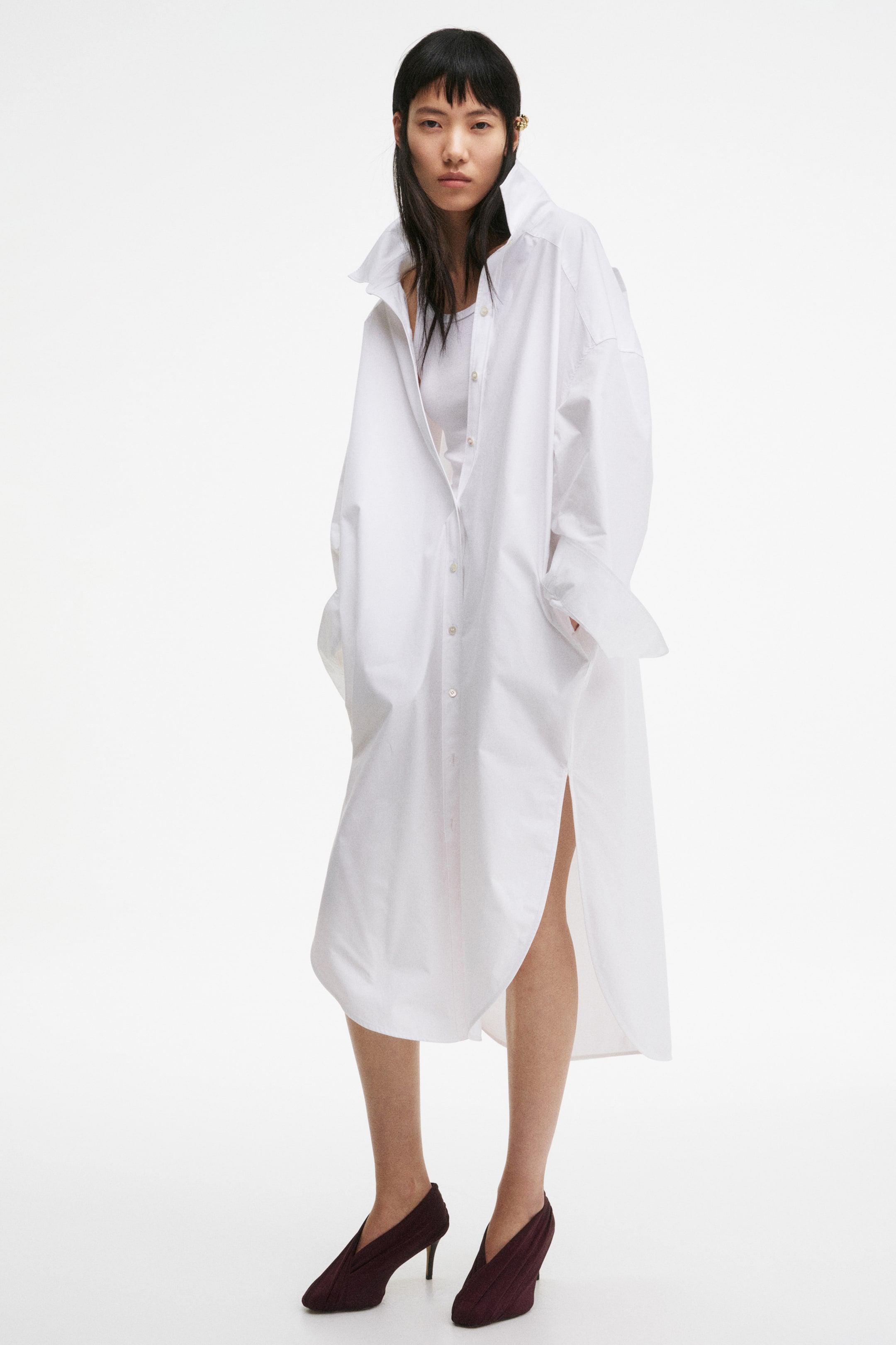 查看大圖: 模特穿著一件小腿長、寬鬆版型的白色襯衫裙,特點包括領子、落肩設計、長寬袖帶袖口,前襟有鈕扣。這件裙子下擺圓弧,側邊有高開衩,模特將雙手放在側口袋中,內搭白色背心。
