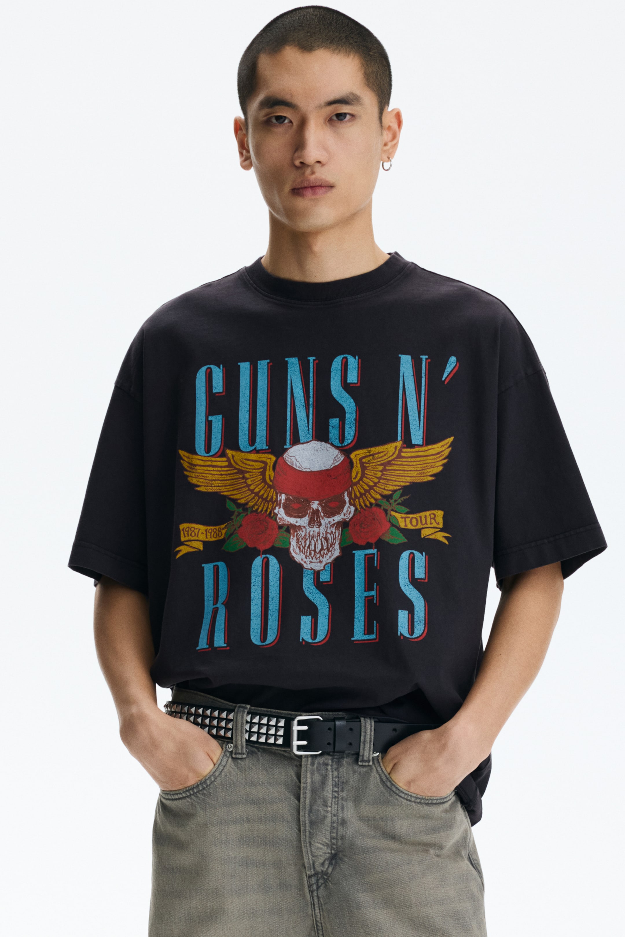 Grotere afbeelding bekijken: Een zwart grafisch T-shirt wordt frontaal getoond, met een vervaagd uiterlijk en een oversized pasvorm. Het heeft een ronde hals en korte mouwen, met een grote centrale afbeelding van een schedel met een rode bandana, gele vleugels en rode rozen, samen met de blauwe tekst "GUNS N' ROSES" en "1987-1988 TOUR."