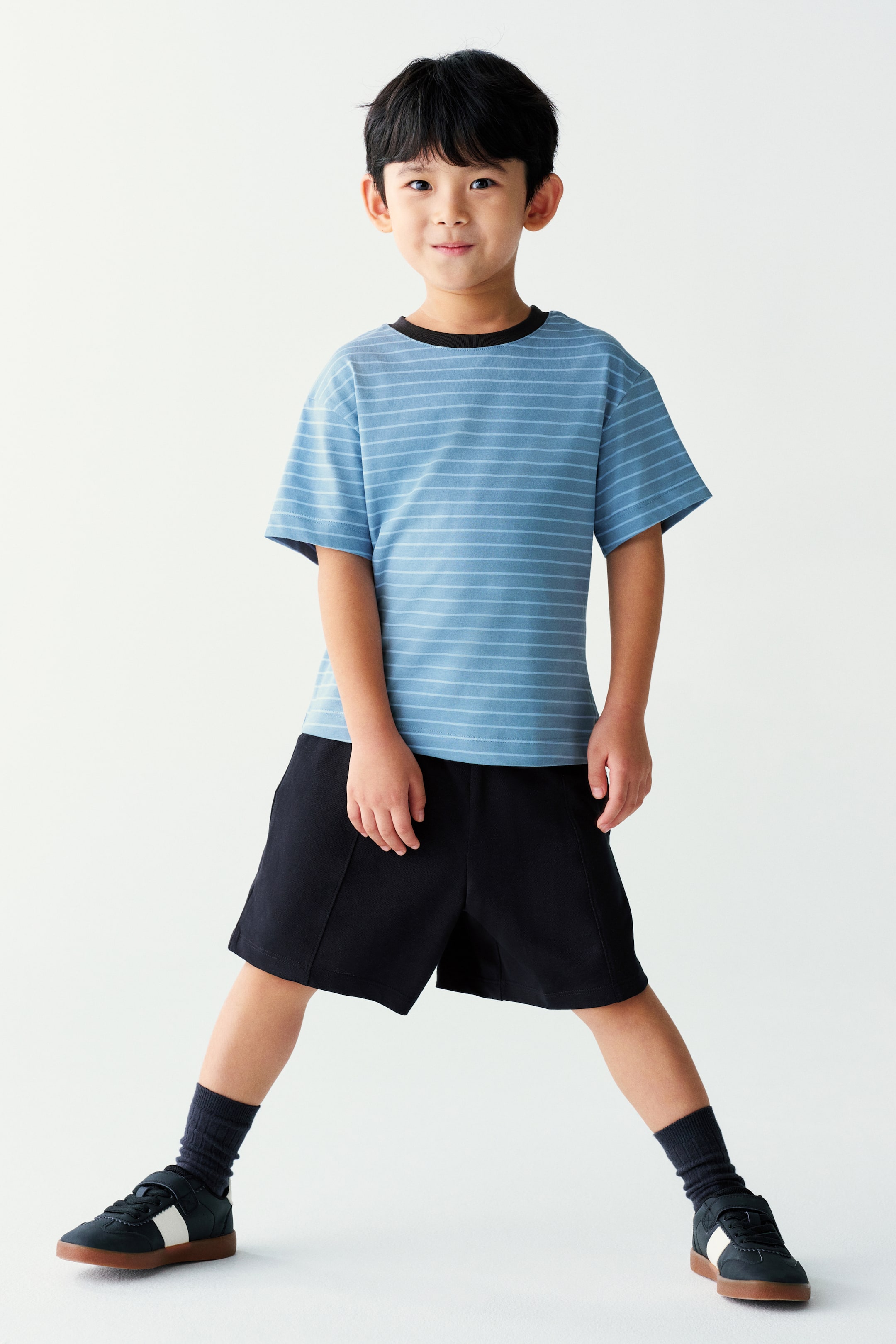 View larger image: A light blue, short-sleeved t-shirt with thin horizontal white stripes and a contrasting black crew neck is being worn by the model. The t-shirt is styled with black shorts, dark socks, and black sneakers.