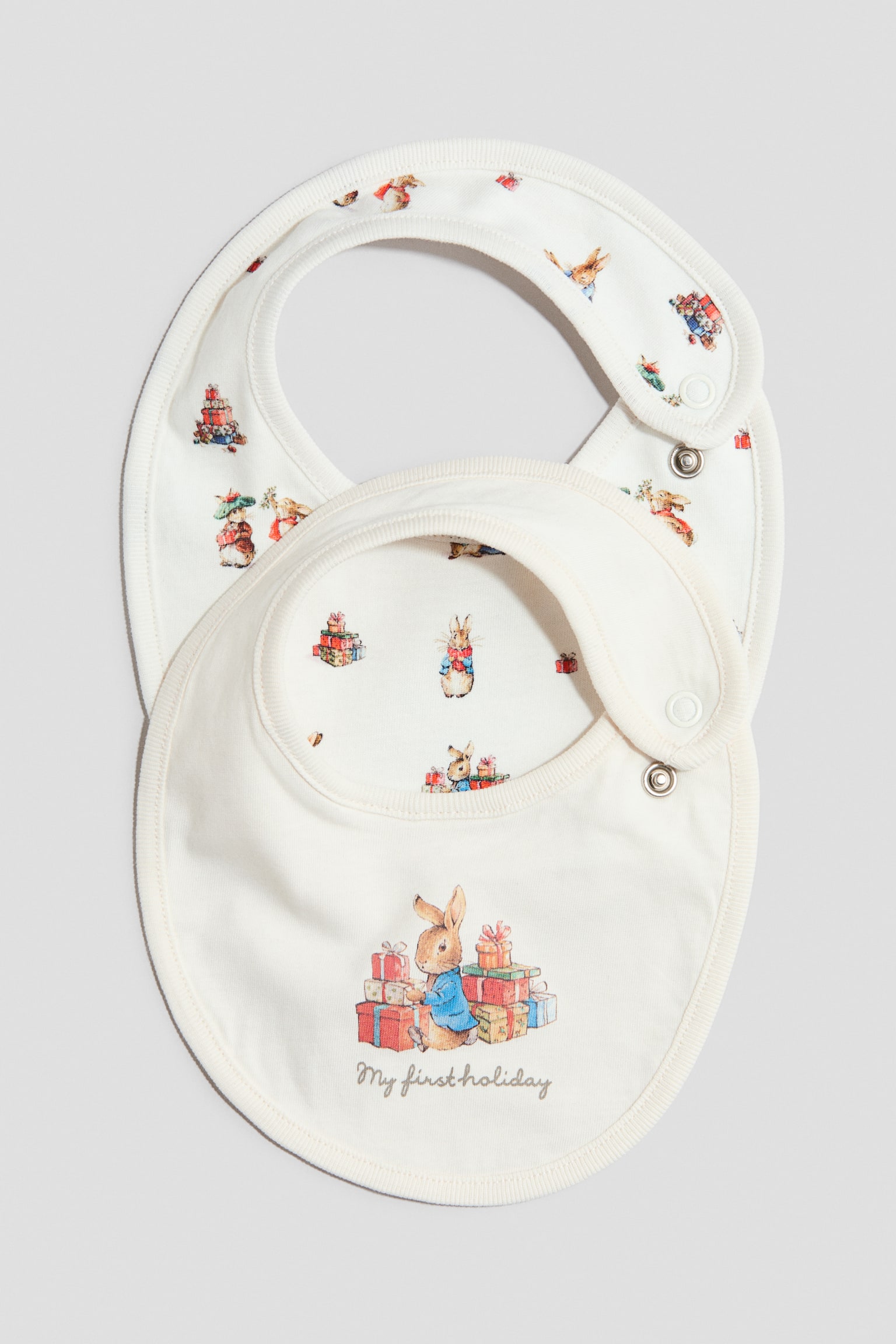 To off-white babyhagesmÃ¦kke vises forfra, hvor den Ã¸verste hagesmÃ¦k har en central illustration af Peter Rabbit ved siden af farverige gaver og teksten "My first holiday" trykt nedenunder. HagesmÃ¦kken nedenunder er med et all-over mÃ¸nster af Peter Rabbit-holidayillustrationer, og begge dele har ribkanter samt trykknaplukning i siden.