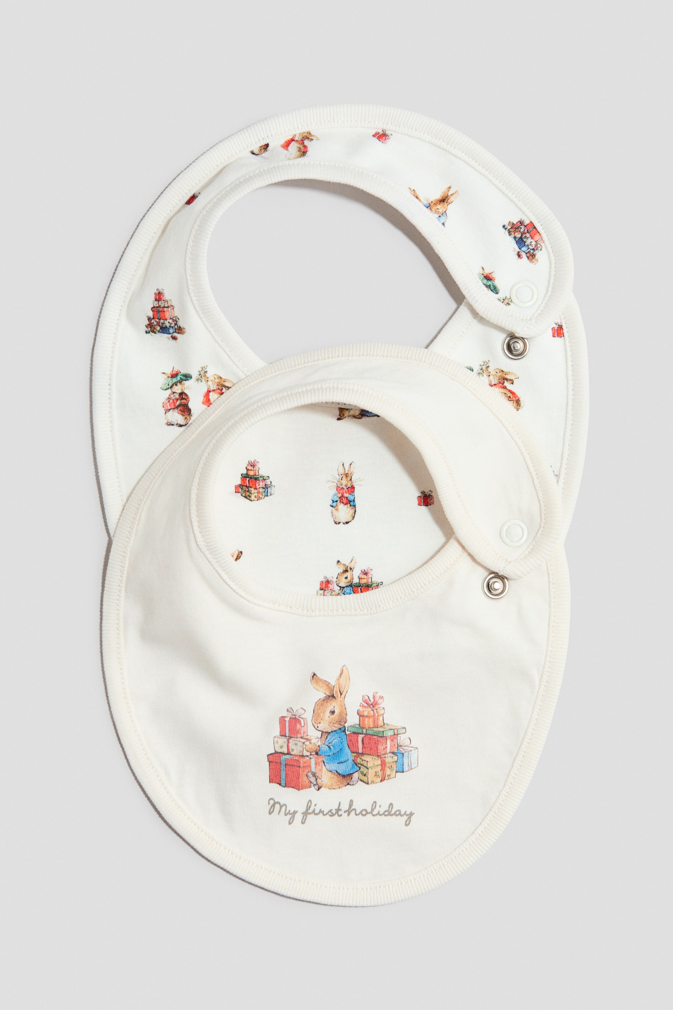 Größeres Bild ansehen: Zwei cremeweiße Baby-Lätzchen sind mit der Vorderseite zu sehen. Das obere Lätzchen zeigt in der Mitte eine Illustration von Peter Rabbit neben farbenfrohen Geschenken, darunter den Aufdruck „My first holiday“. Das darunterliegende Lätzchen ist mit einem Allover-Muster aus Peter-Rabbit-Weihnachtsillustrationen versehen. Beide Modelle verfügen über gerippte Abschlüsse und seitliche Druckknopfverschlüsse.