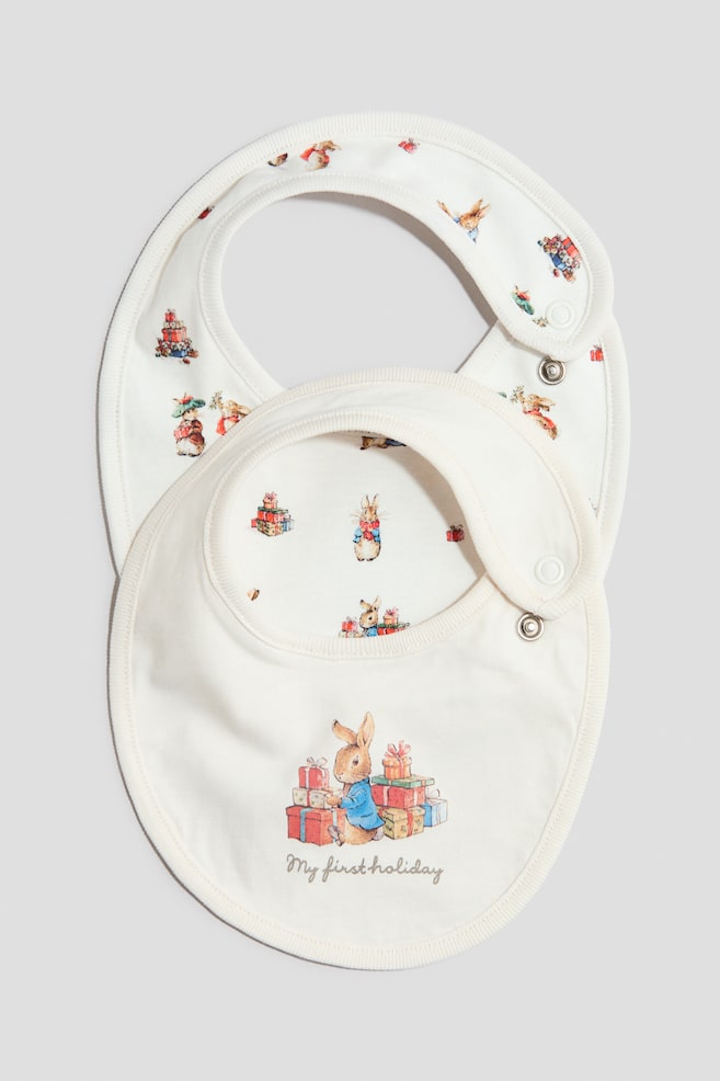Deux bavoirs pour bÃ©bÃ© blanc cassÃ© sont prÃ©sentÃ©s de face : celui du dessus affiche une illustration centrale de Peter Rabbit Ã  cÃ´tÃ© de cadeaux colorÃ©s, avec le texte Â« My first holiday Â» imprimÃ© en dessous. Le bavoir en dessous prÃ©sente un motif intÃ©gral dâillustrations de vacances de Peter Rabbit, les deux piÃ¨ces mettant en valeur des bordures cÃ´telÃ©es et des fermetures Ã  boutons-pression sur le cÃ´tÃ©. - 1