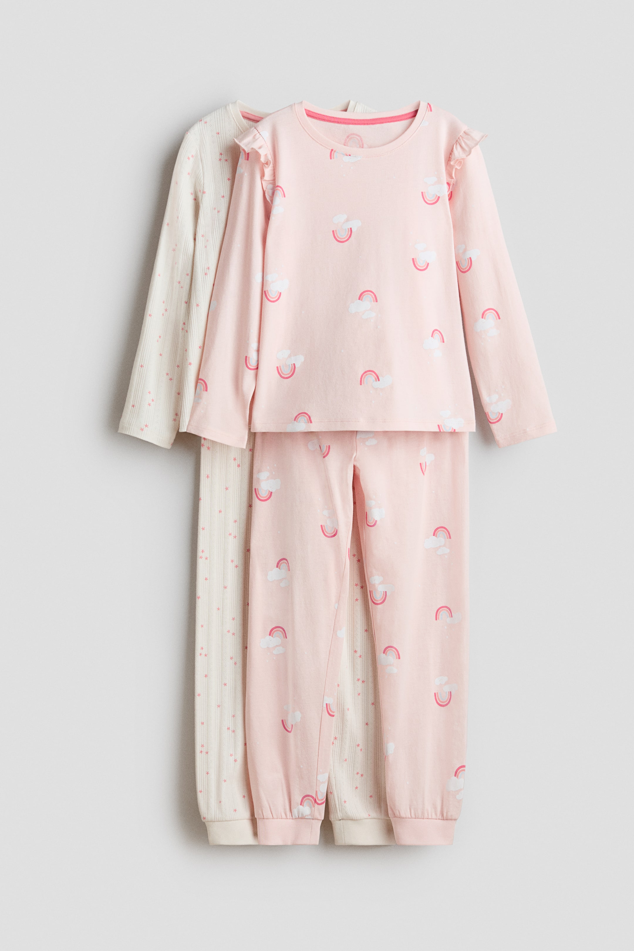 View larger image: Presenting two sets of pyjamas, each comprising a top and bottom crafted from soft, printed cotton jersey. The front set in pink showcases a long-sleeved top with a round neckline and ruffled shoulders, adorned with prints of white clouds and pink rainbows, complemented by matching bottoms featuring ribbed hems. Layered behind is a cream-colored set, distinguished by a vertically ribbed long-sleeved top and bottoms embellished with small pink star prints.