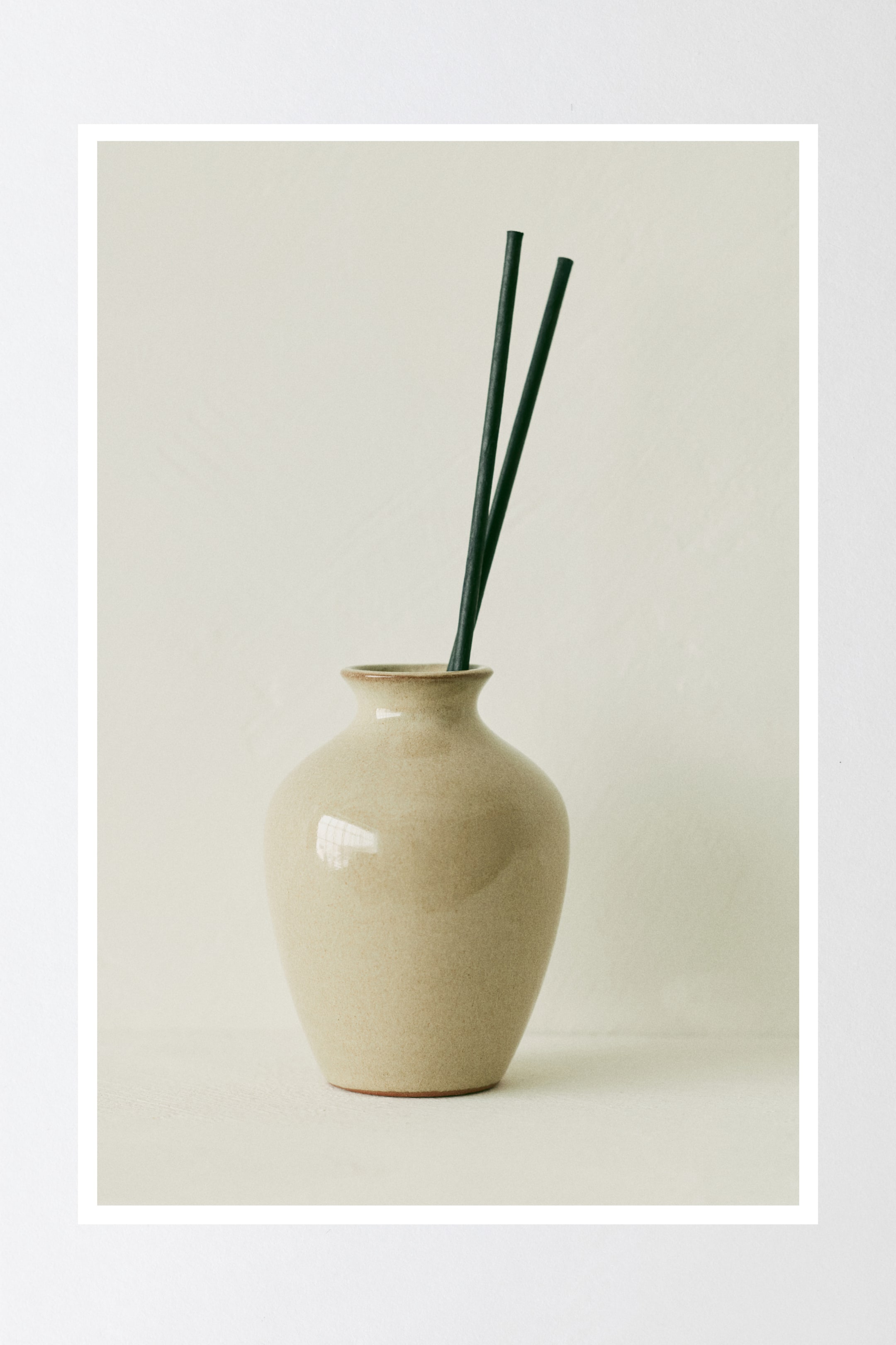 Grösseres Bild ansehen: Eine hellbeige, glänzende Keramik-Diffusorvase mit einem bauchigen Körper und einem schmalen, ausgestellten Rand hält zwei dunkle Rattanstäbe, die aufrecht stehen und leicht geneigt sind.