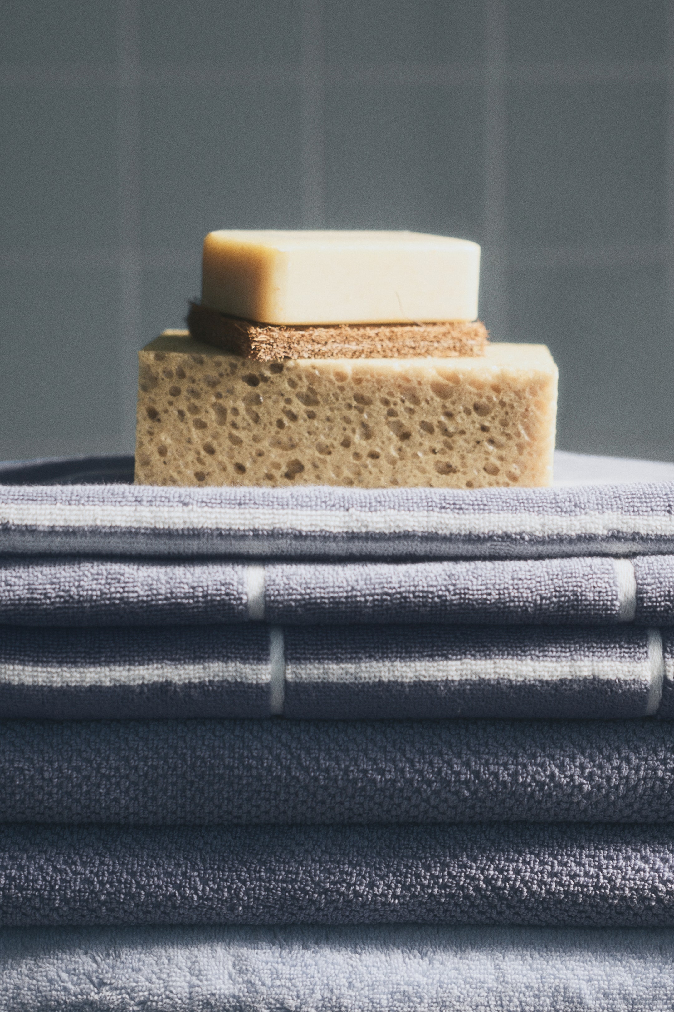 View larger image: A stack of soft cotton terry bath towels is displayed, neatly folded. The top towels feature an accent-colour grid pattern, with white horizontal lines visible on a grey-blue background. The lower towels display a solid grey-blue colour and their terry texture.