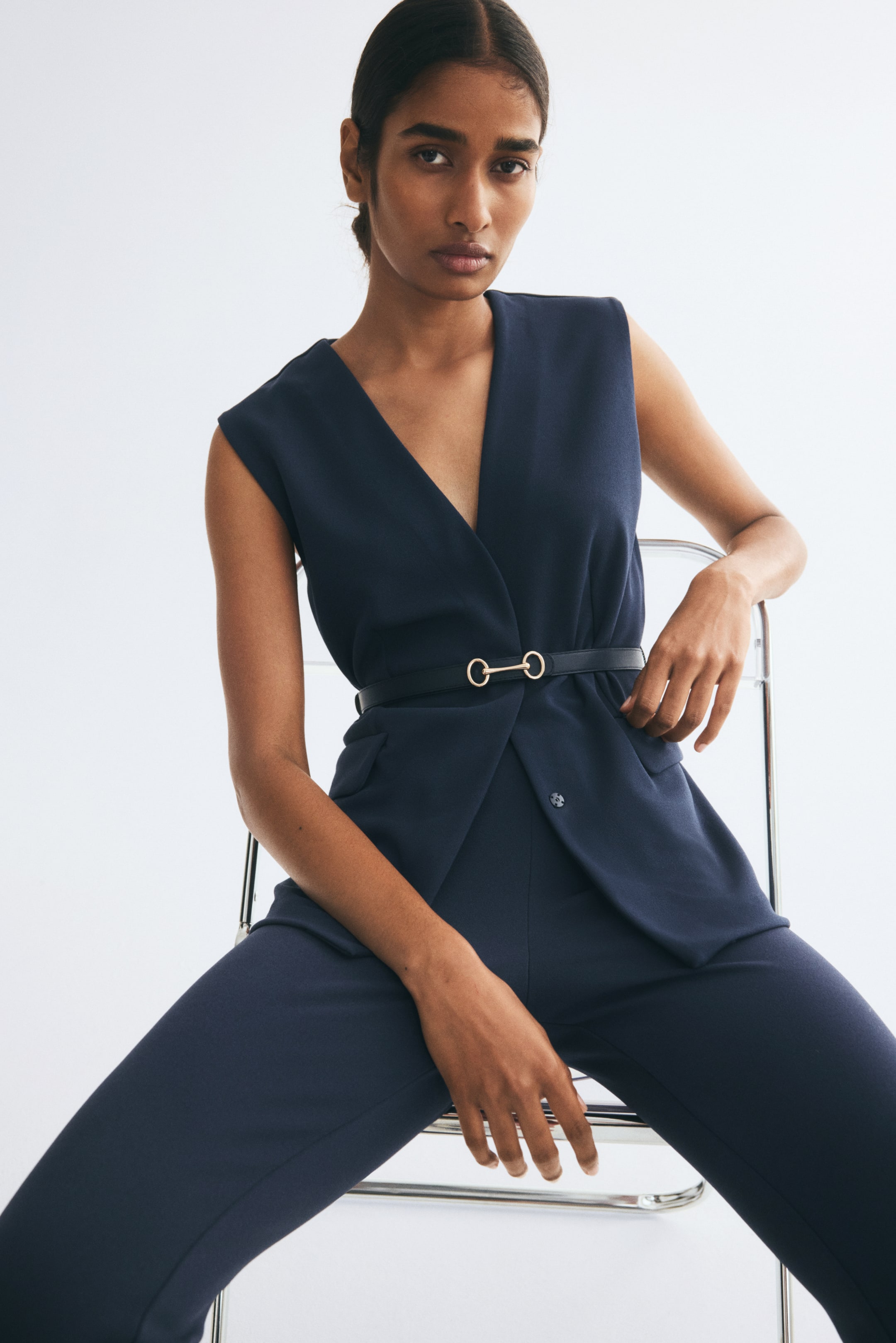 View larger image: The model is wearing a navy blue vest in stretch jersey, paired with matching trousers. The vest features a deep V-neckline, decorative side pocket flaps, and a slim black waist belt with a gold-toned metal buckle.