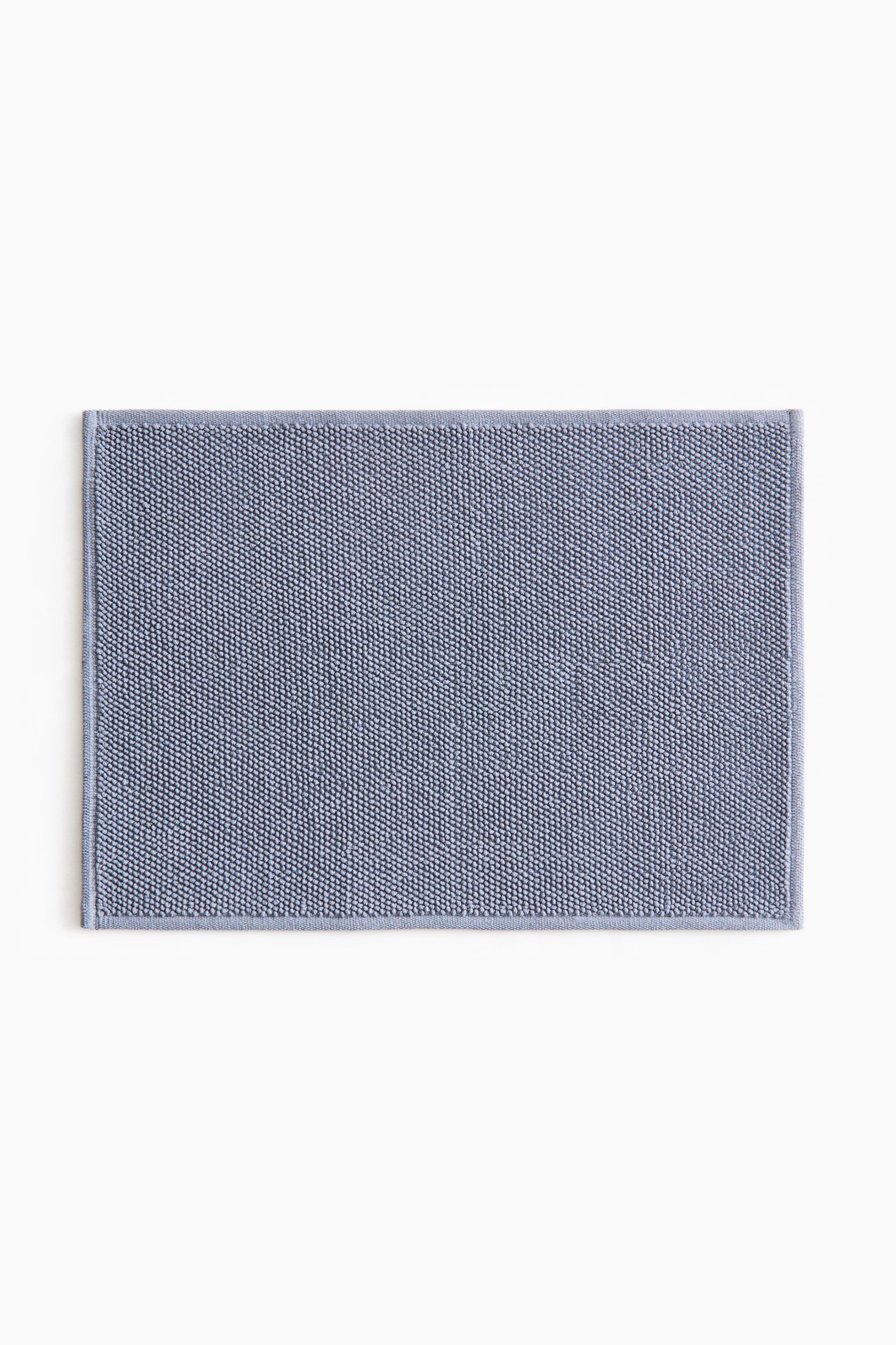 View larger image: A rectangular blue-grey bath mat showcases its textured surface, adorned with small, dense loops. The mat is completed with a flat, woven border along its edges.